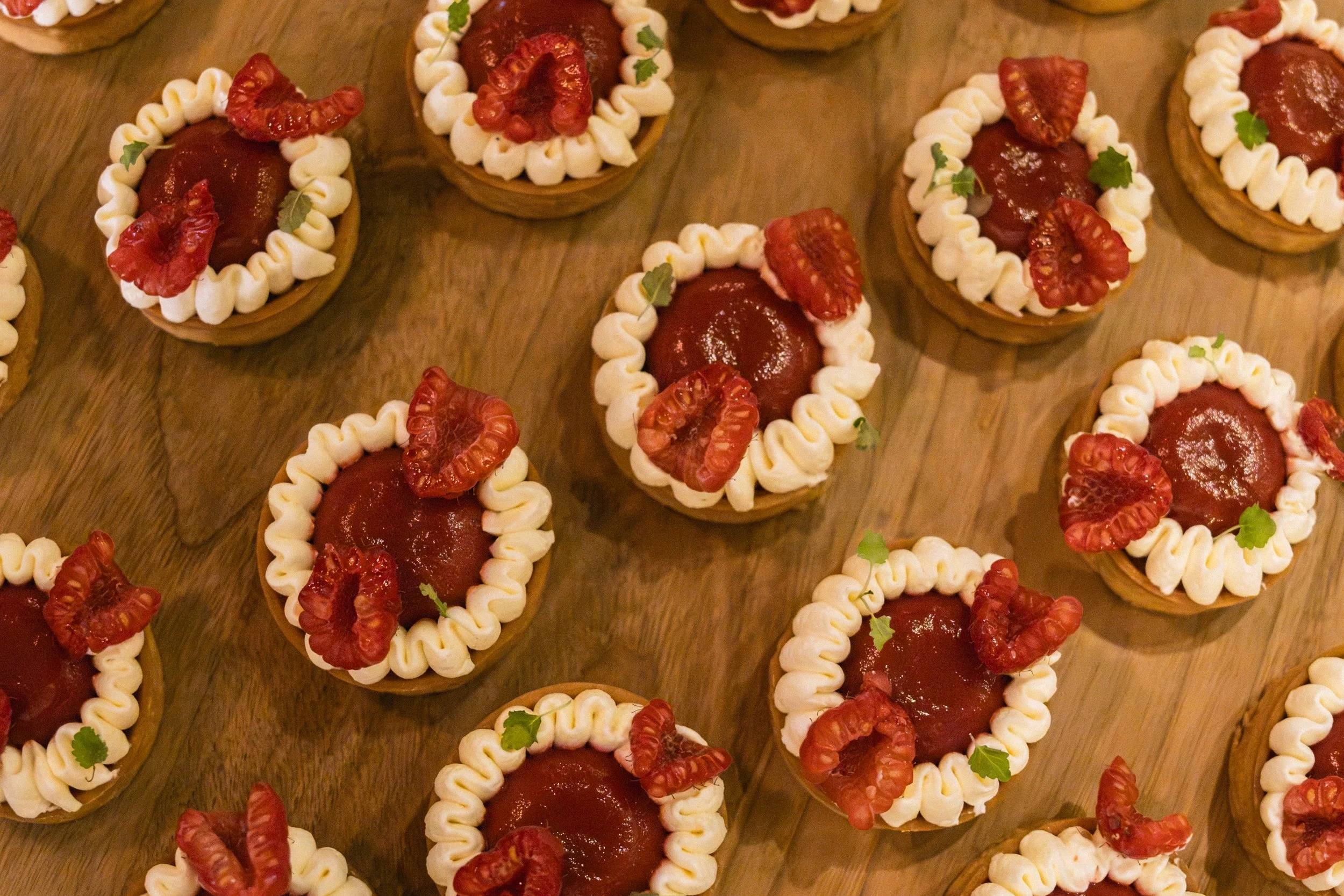 Multiple individual tartlets with cream border, topped with red fruit jam, dried red fruit, and small green herbs on a wooden surface.