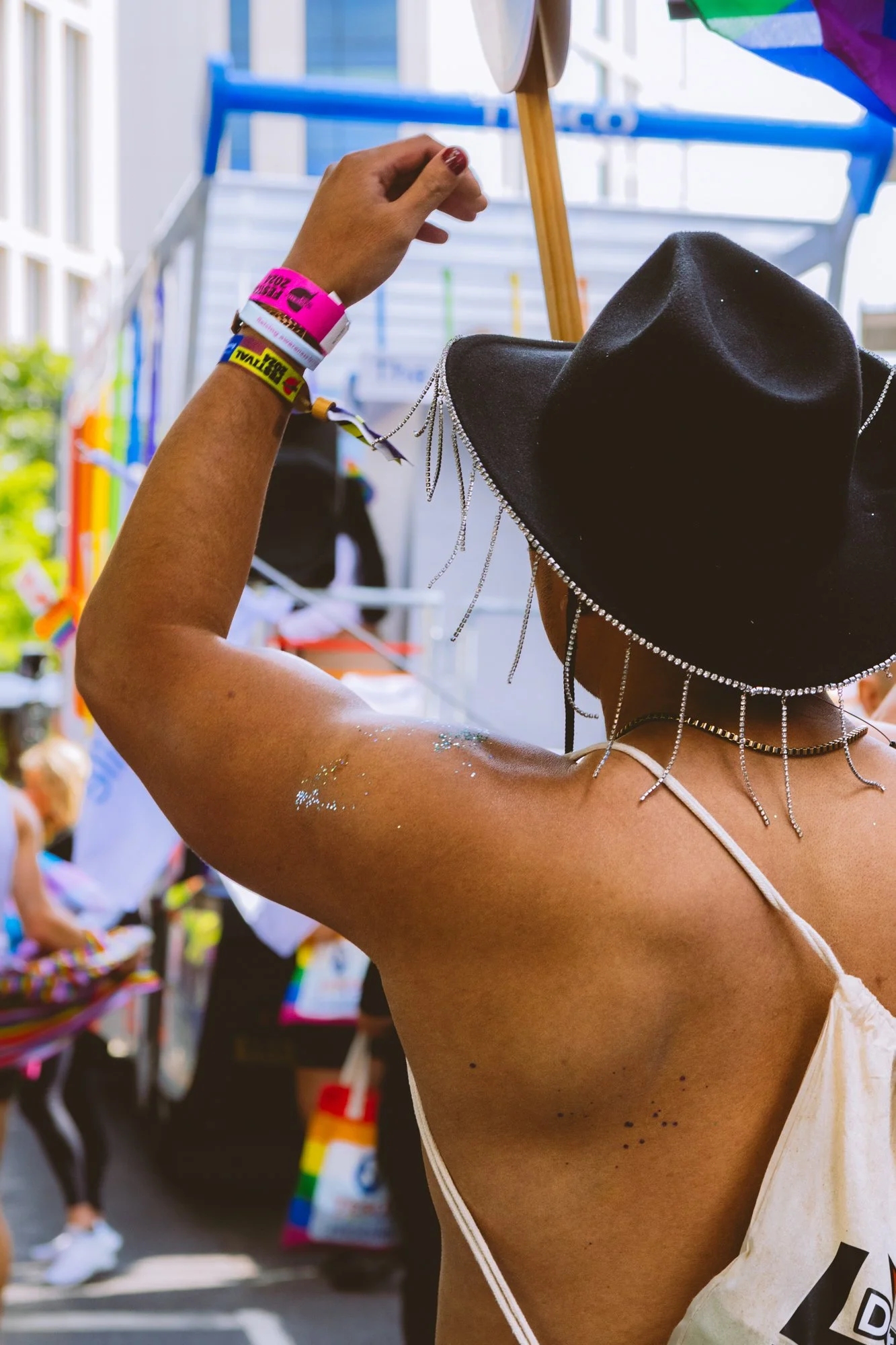 A person wearing a black hat with silver chains and a sleeveless top, with glitter on their arm, is reaching for items at an outdoor event or marketplace.