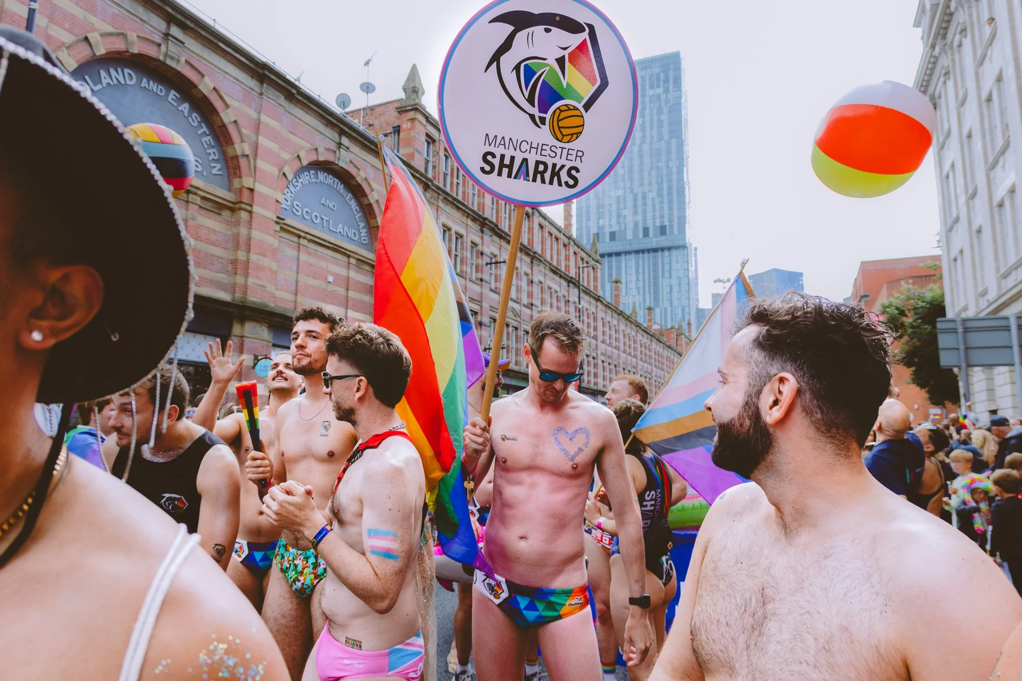 A group of people participating in a pride parade in Manchester, dressed in colorful underwear, holding rainbow flags and rainbow-colored balloons. Some individuals are shirtless and celebrating in the street, with historic buildings in the backgroun