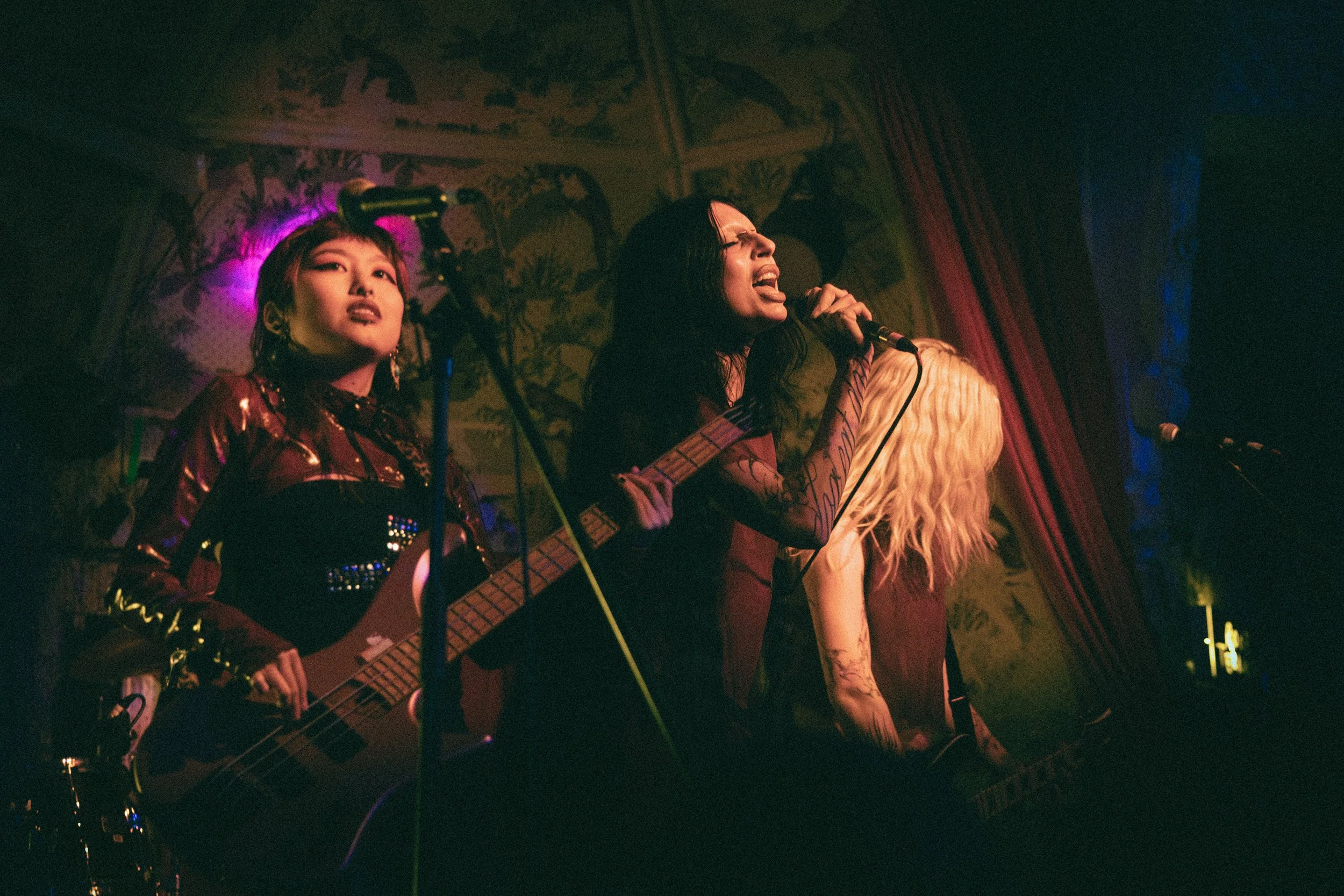 Three performers singing and playing guitar on stage with dark, ornate wallpaper and red curtains behind them, under colourful stage lighting.