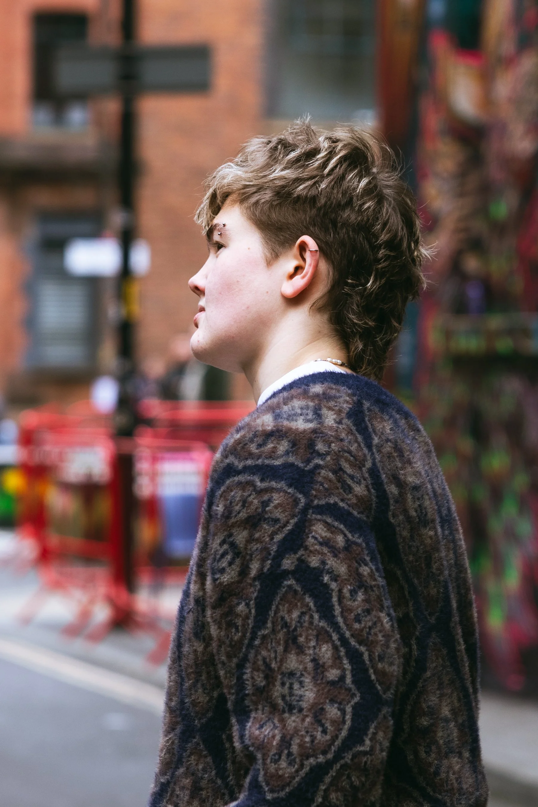 Side profile of a person with short, wavy hair, standing in the street with red road work fencing in the background.