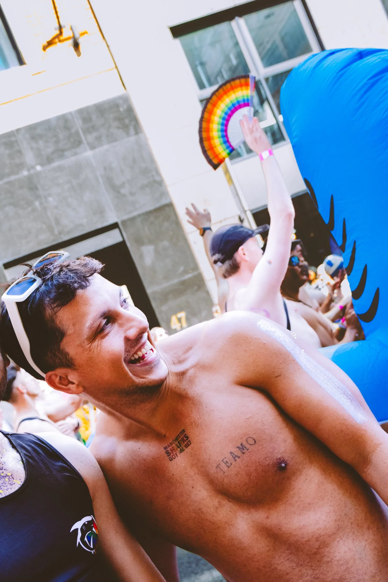 A group of people at a summer outdoor event, with a man smiling and shirtless in the foreground. Others in the background are holding colorful fans and wearing casual summer attire, with a sunlit building behind them.