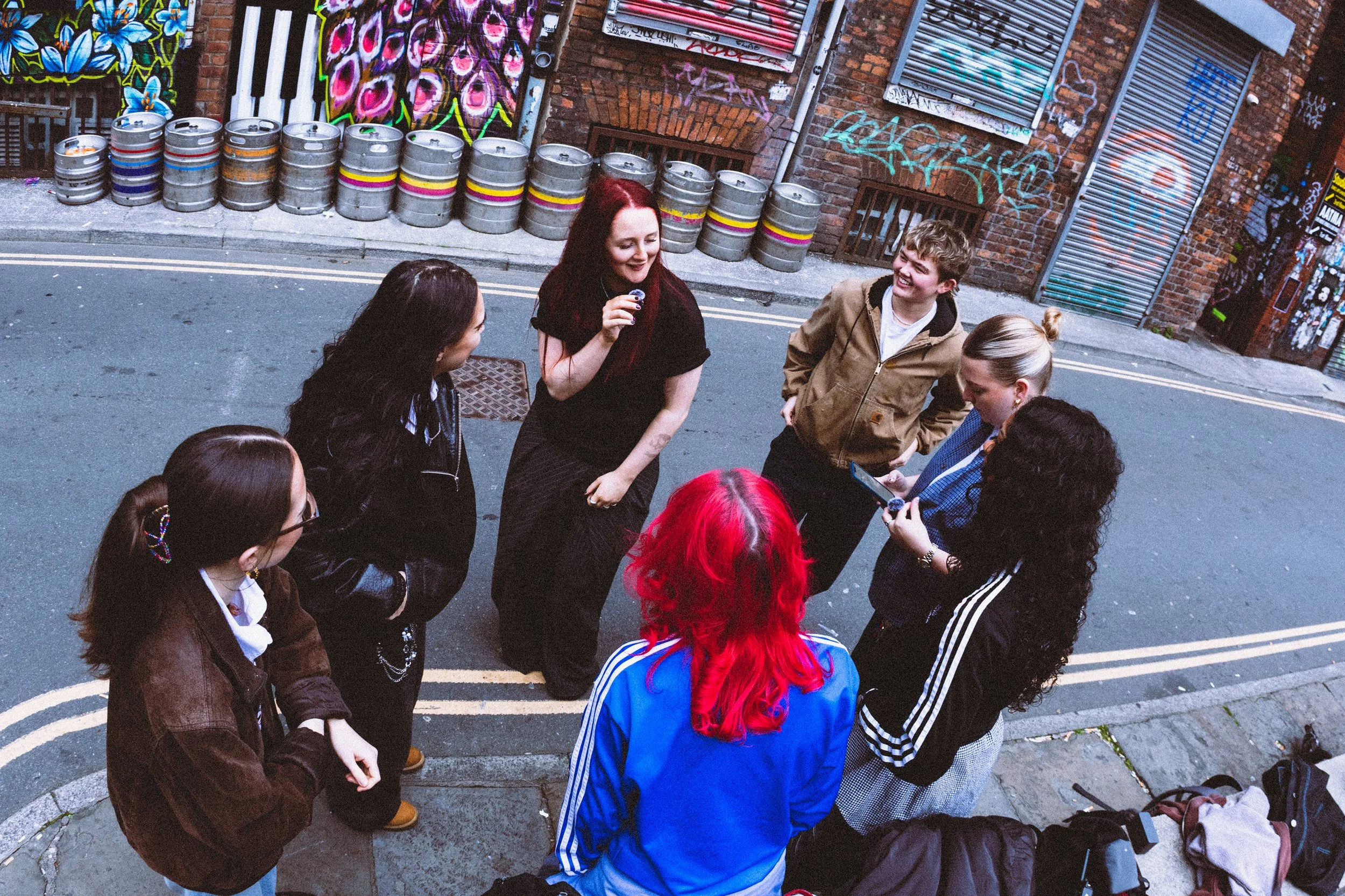 A group of young people standing in a circle and talking on a street with brick buildings in the background, surrounded by graffiti art and beer kegs.