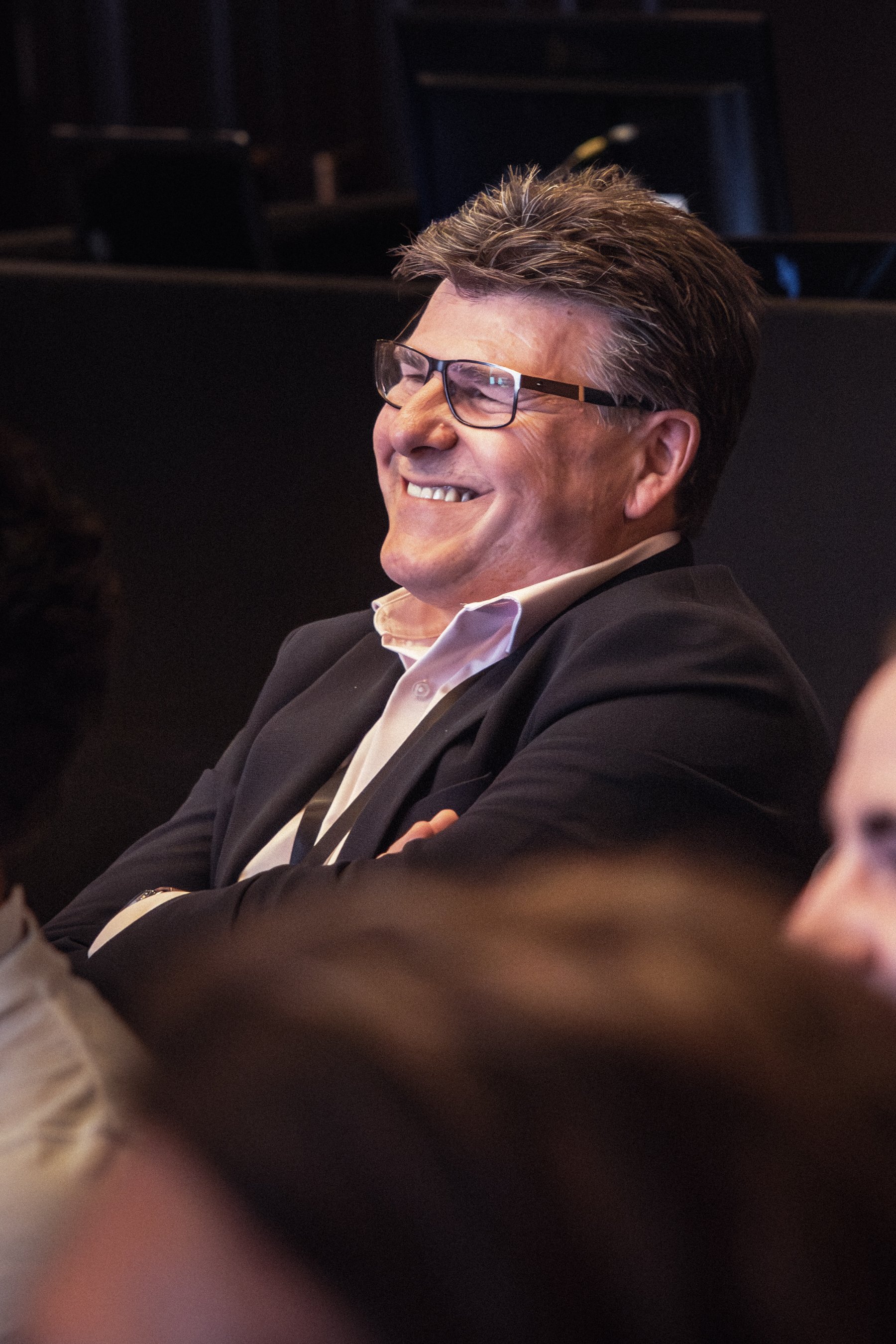 A man with dark hair, glasses, and a suit, smiling and laughing at a conference.