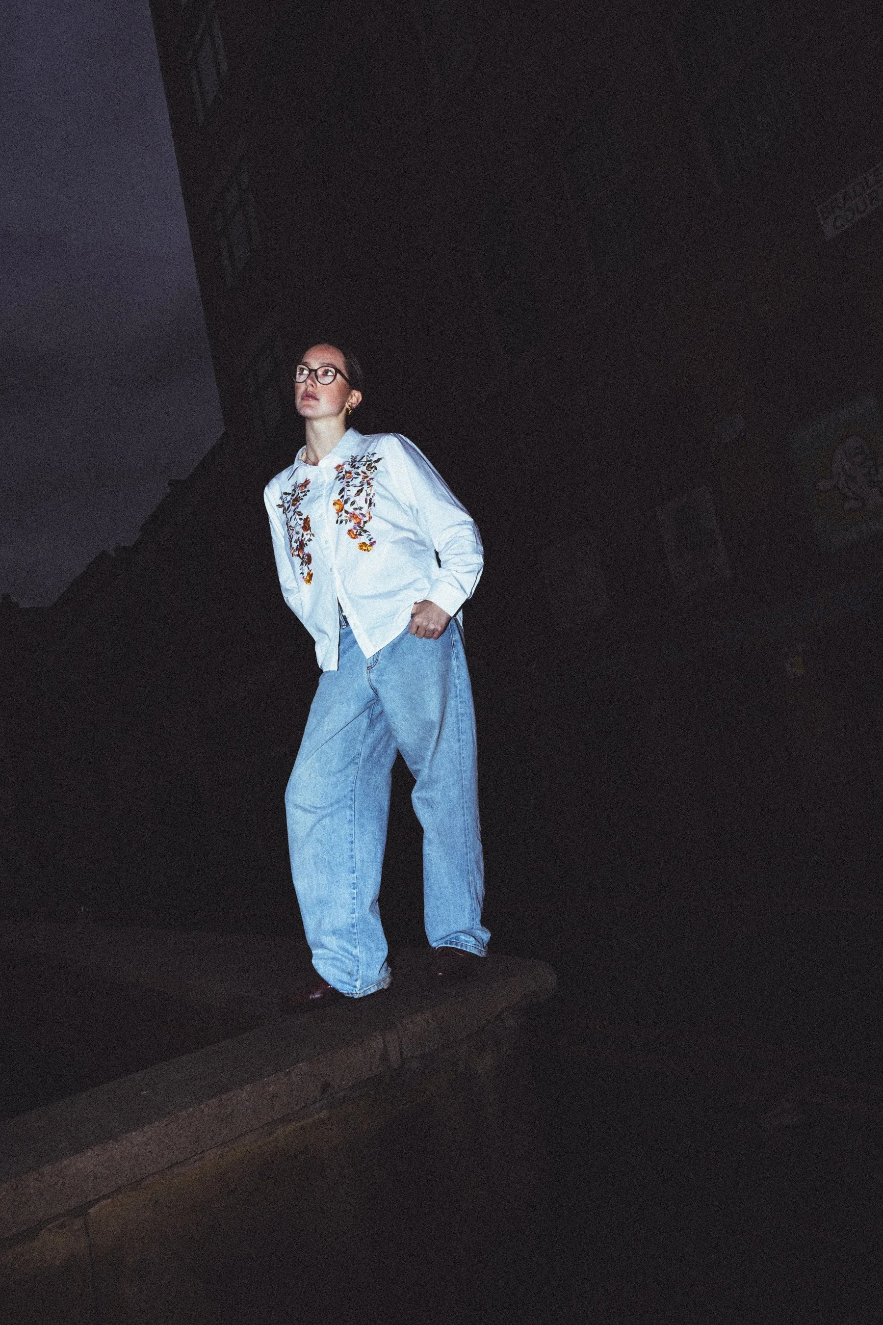 A person standing outdoors on a wall, wearing glasses, a white embroidered shirt, and baggy light blue jeans.