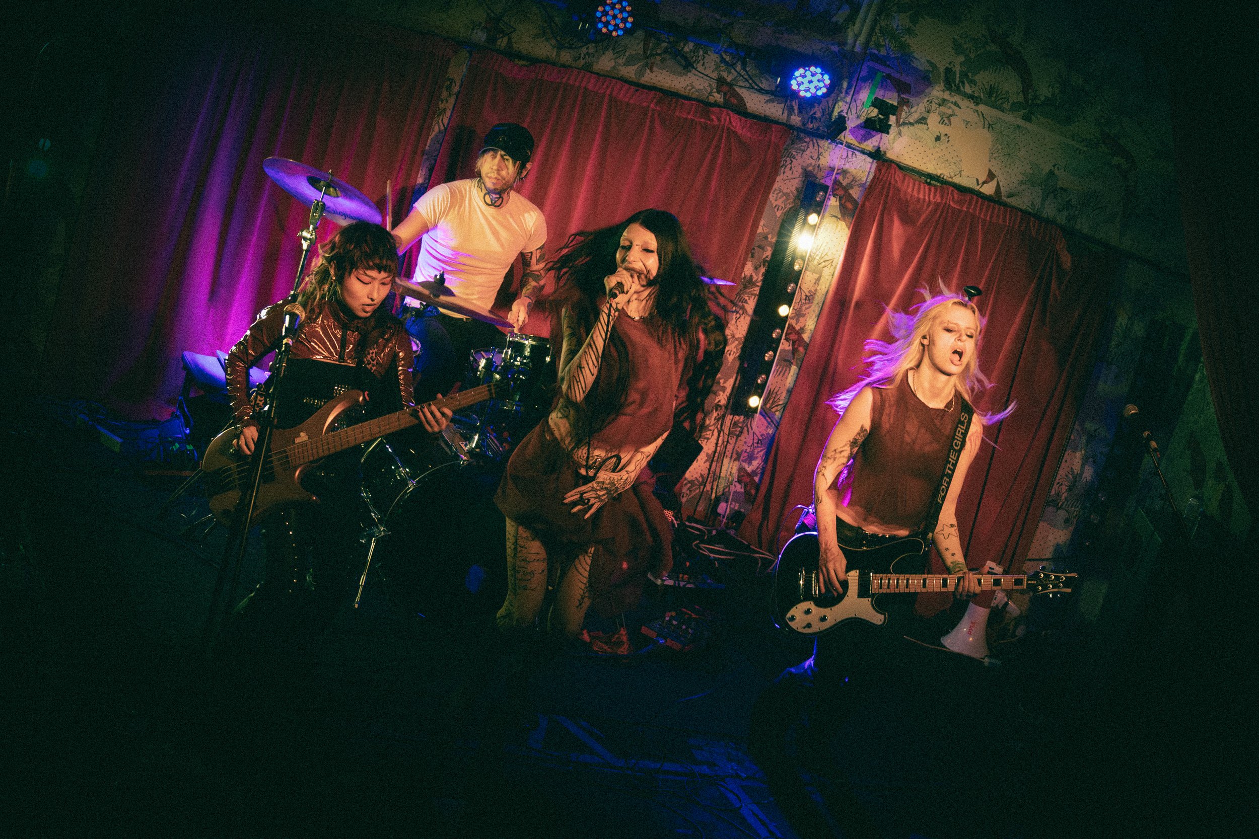 Four musicians performing on a stage with red curtains and colorful lights. Two in the front, one singing into a microphone and the other playing an electric guitar. Behind them, a man plays the drums.