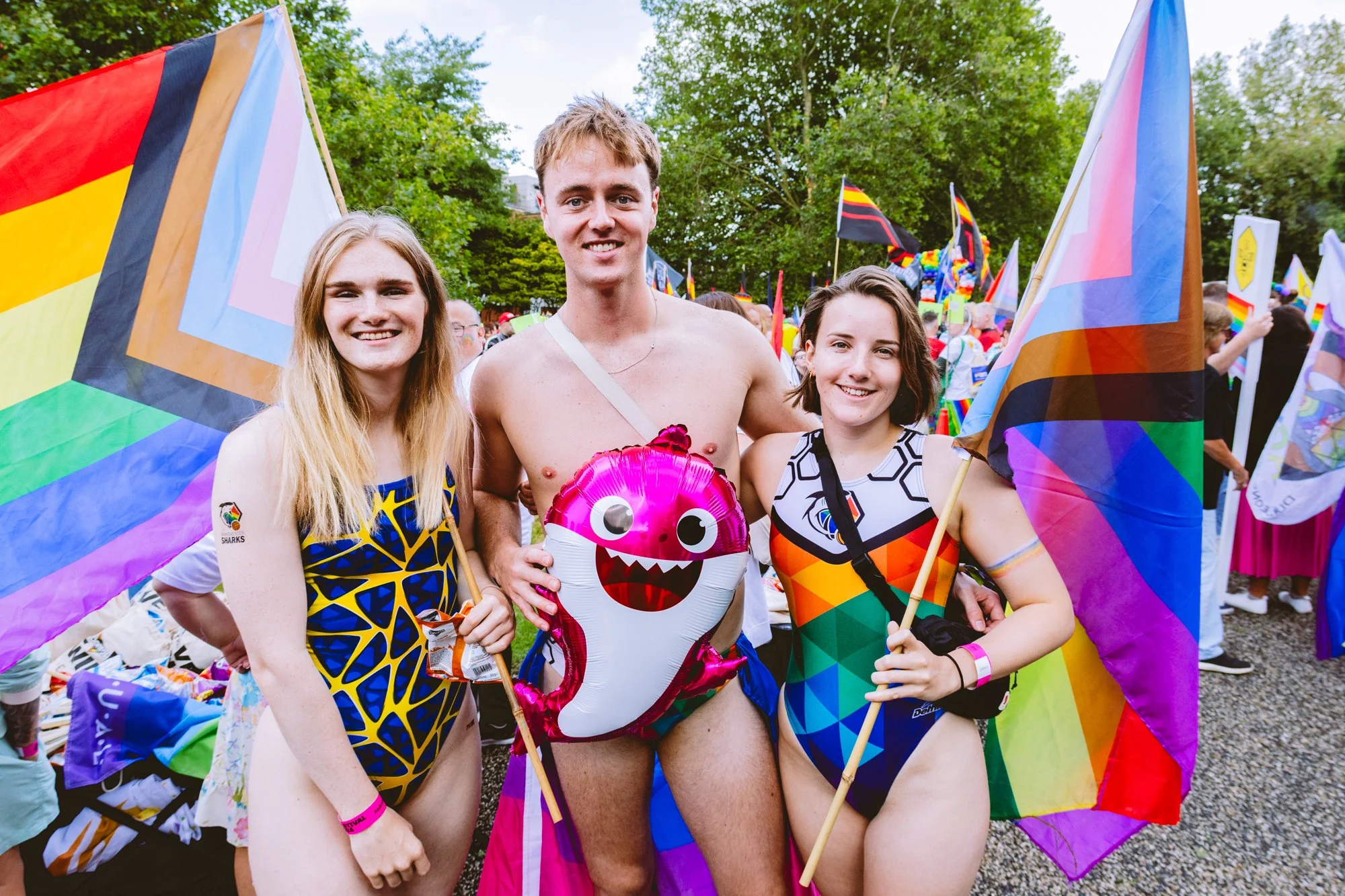 Three smiling individuals at an outdoor pride event, holding rainbow flags and a pink heart-shaped foil balloon with a cartoon monster face. One person is shirtless, flanked by two women. The background shows a crowd with flags and trees.