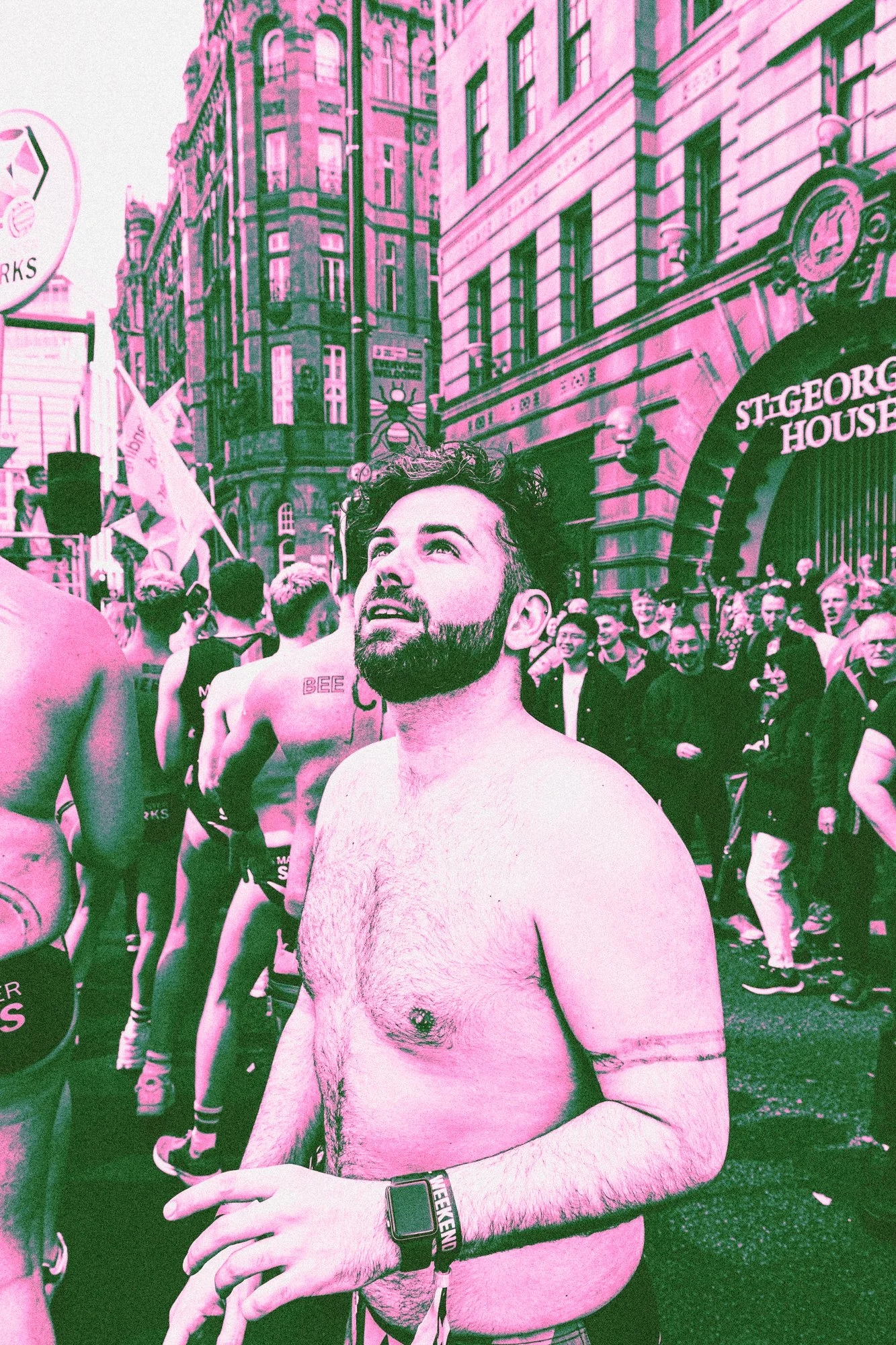 A shirtless man with a beard and short hair standing outdoors during Manchester Pride. Behind him, many people are gathered, some holding flags and banners.