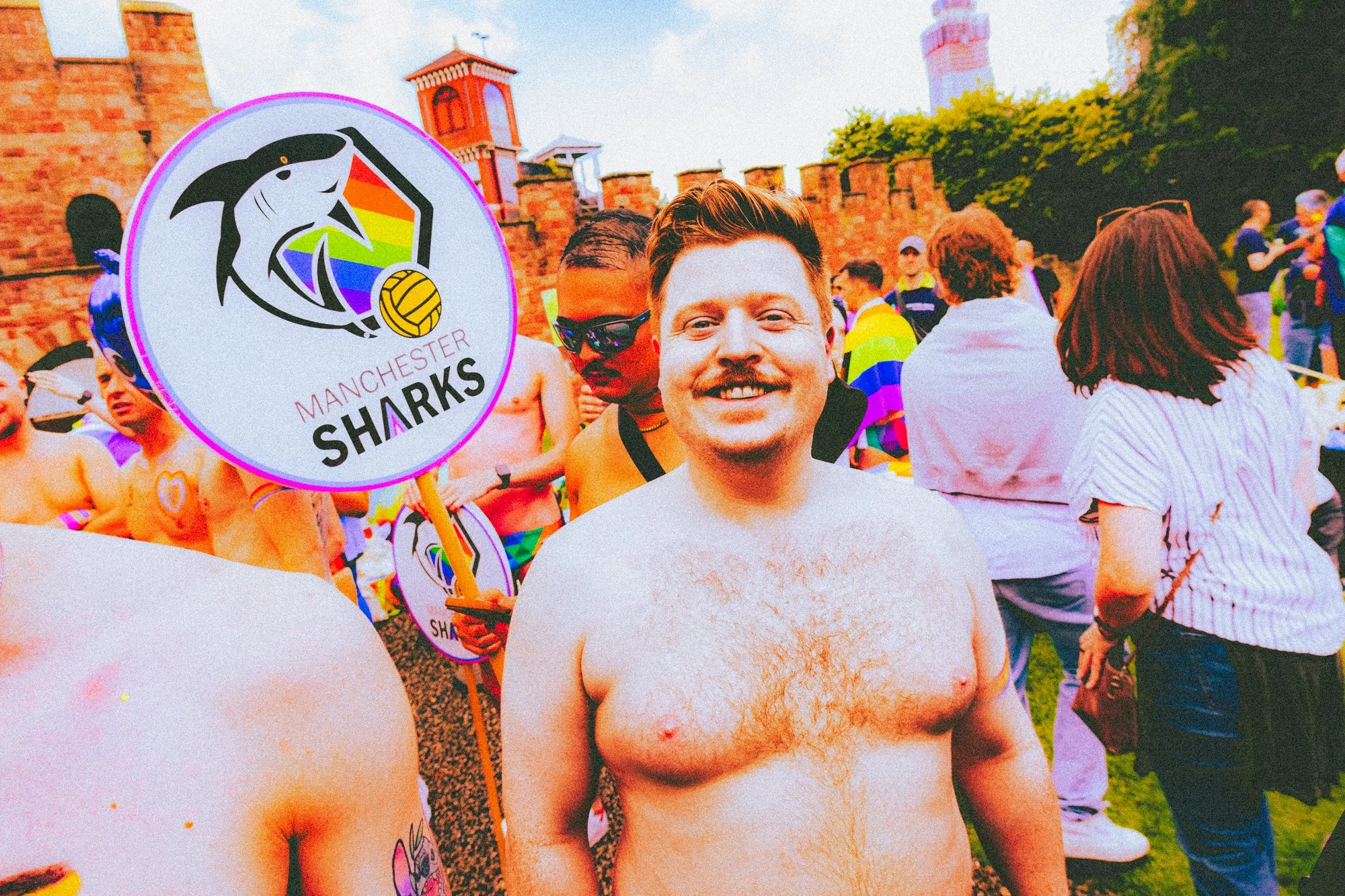 A group of people participating in a pride event, with some holding signs for the Manchester Sharks, a volleyball team. The main focus is on a shirtless man with a beard and mustache, smiling at the camera. Other participants are wearing rainbow flag