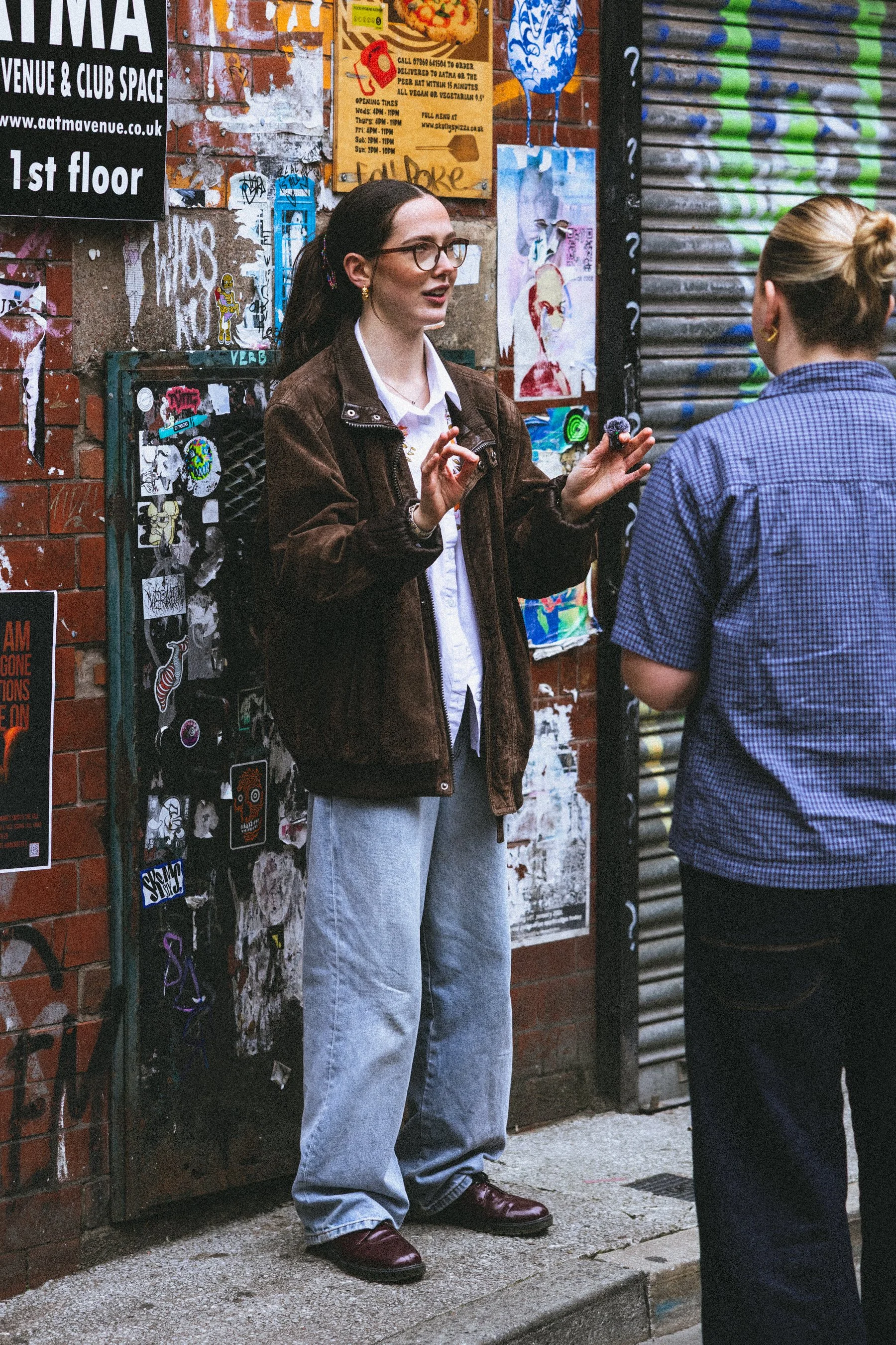Two people having a conversation on a city sidewalk. One has long dark hair, glasses, a brown jacket, and light blue jeans. The other has blonde hair tied back, earrings, and a checked shirt.