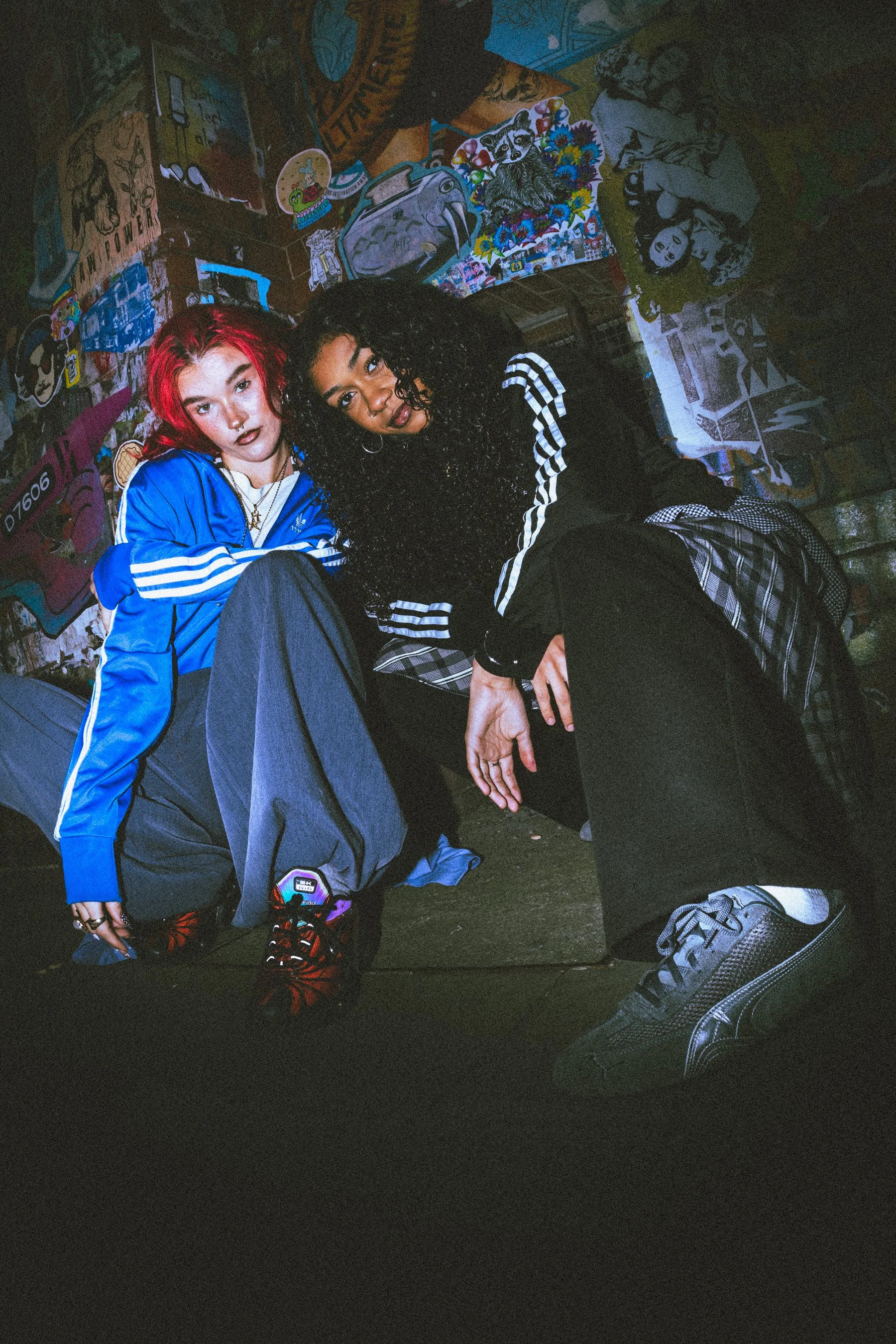 Two young women sitting on the ground surrounded by artwork and posters on a wall, one with bright red hair and the other with curly black hair, both wearing athletic clothing.