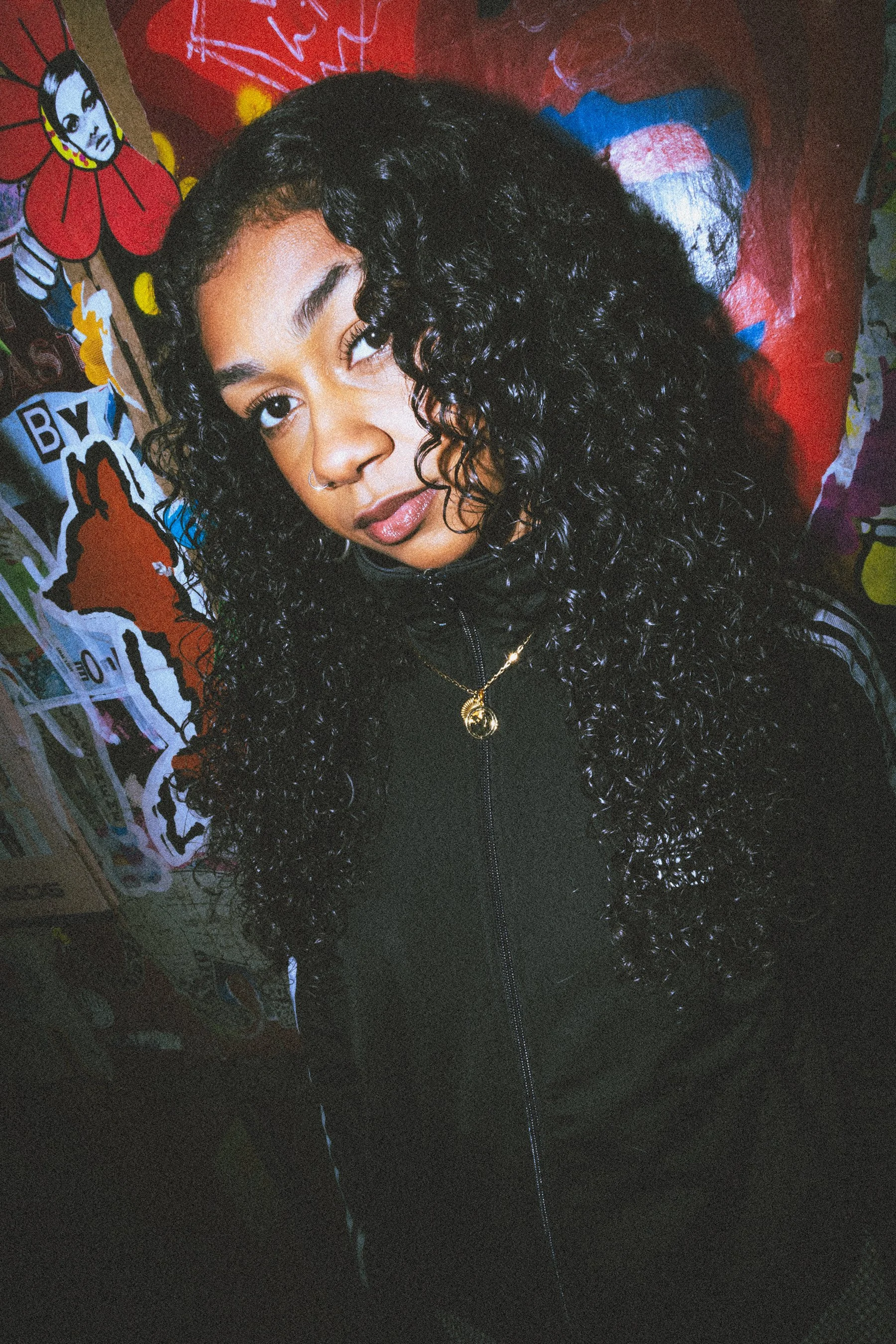 Close-up of a person with dark curly hair, wearing a black top and gold necklace, standing in front of colourful graffiti art.