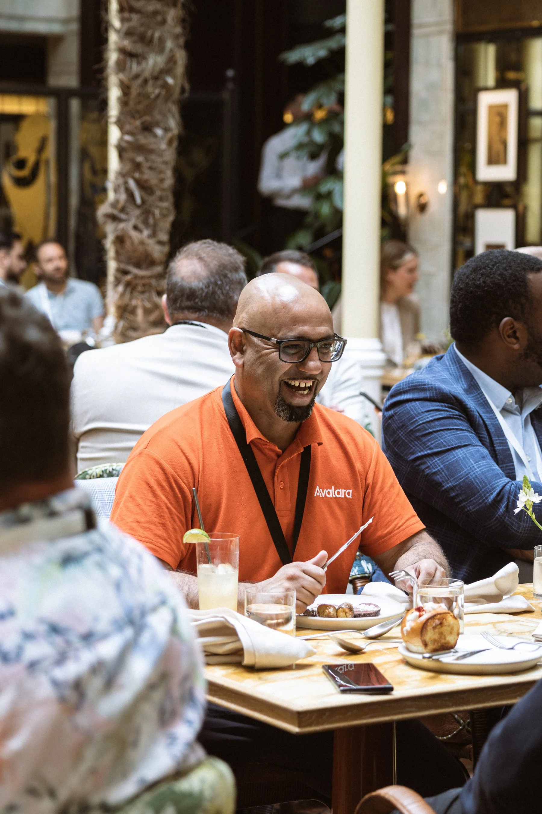 People sitting around a table enjoying a meal in a cozy restaurant with wood and plant decor. A man in an orange shirt with glasses is smiling, while others are engaged in conversation.