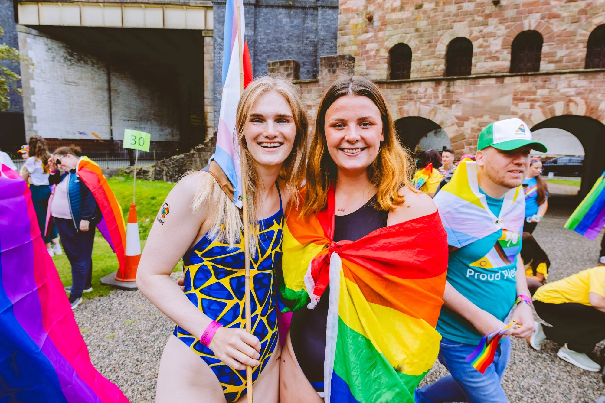 Two smiling women at a pride parade, one wearing a rainbow patterned outfit and the other wrapped in a rainbow pride flag, standing in front of a brick building with a crowd of people celebrating.