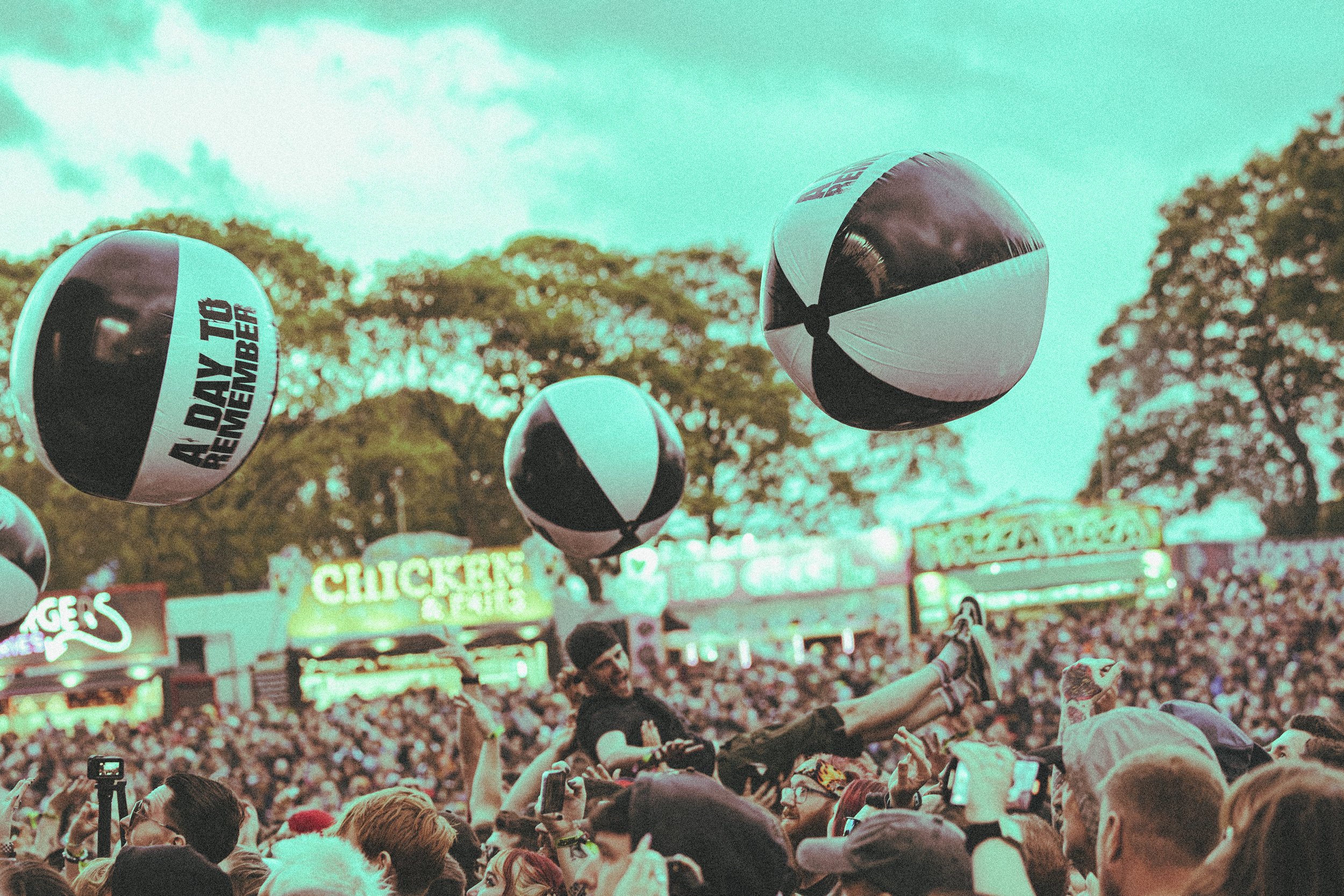Crowd at Slam Dunk festival with floating beach balls marked 'A DAY TO REMEMBER' in the air, several booths with signs, and trees in the background.