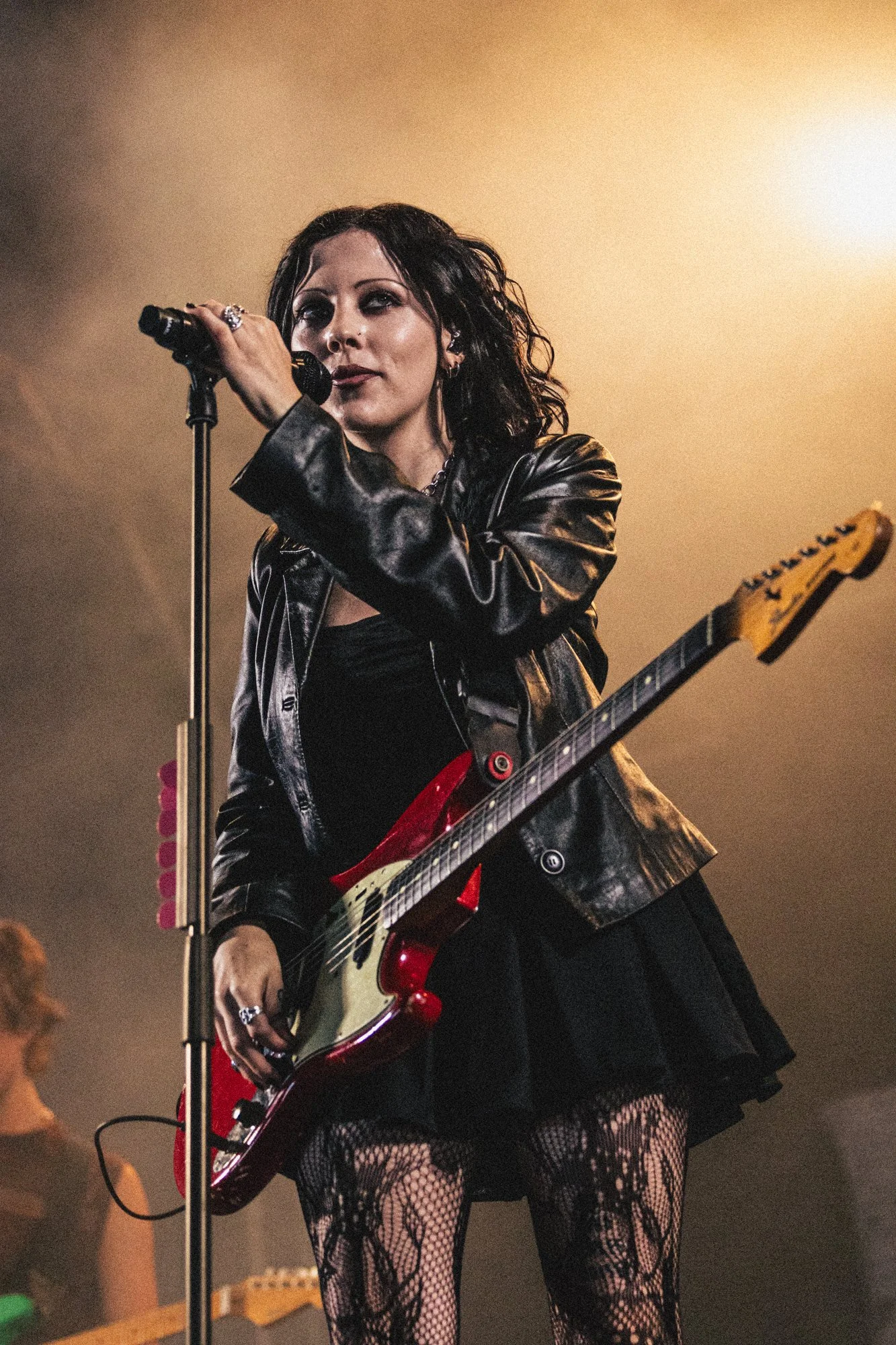 A female musician performs on stage holding a microphone and a red electric guitar, wearing a leather jacket and fishnet stockings.