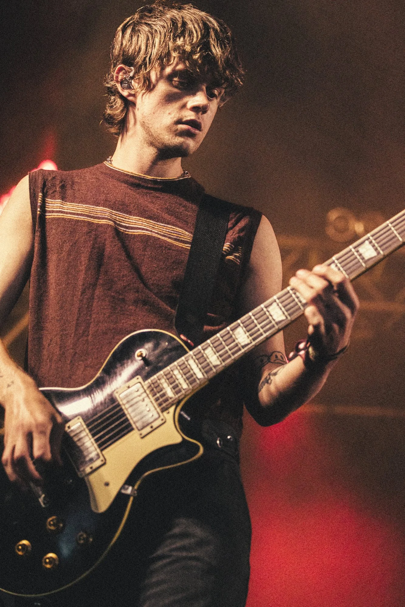A young man with curly brown hair playing an electric guitar on stage with a dark background and red lighting.