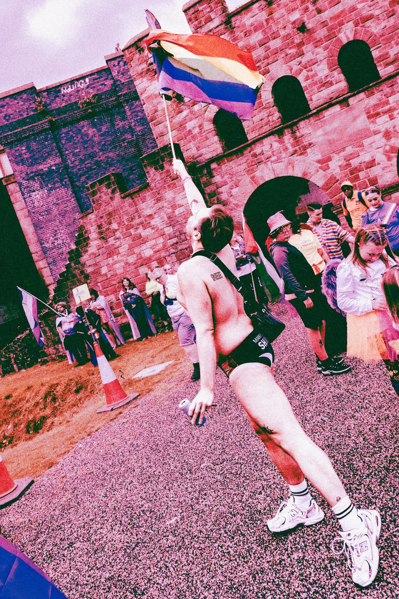 Person with short hair wearing sunglasses, a tank top, shorts, and sneakers, holding a drink in one hand, walking on a gravel path in front of a historic brick castle with arched windows, surrounded by a group of people, some holding rainbow flags, a