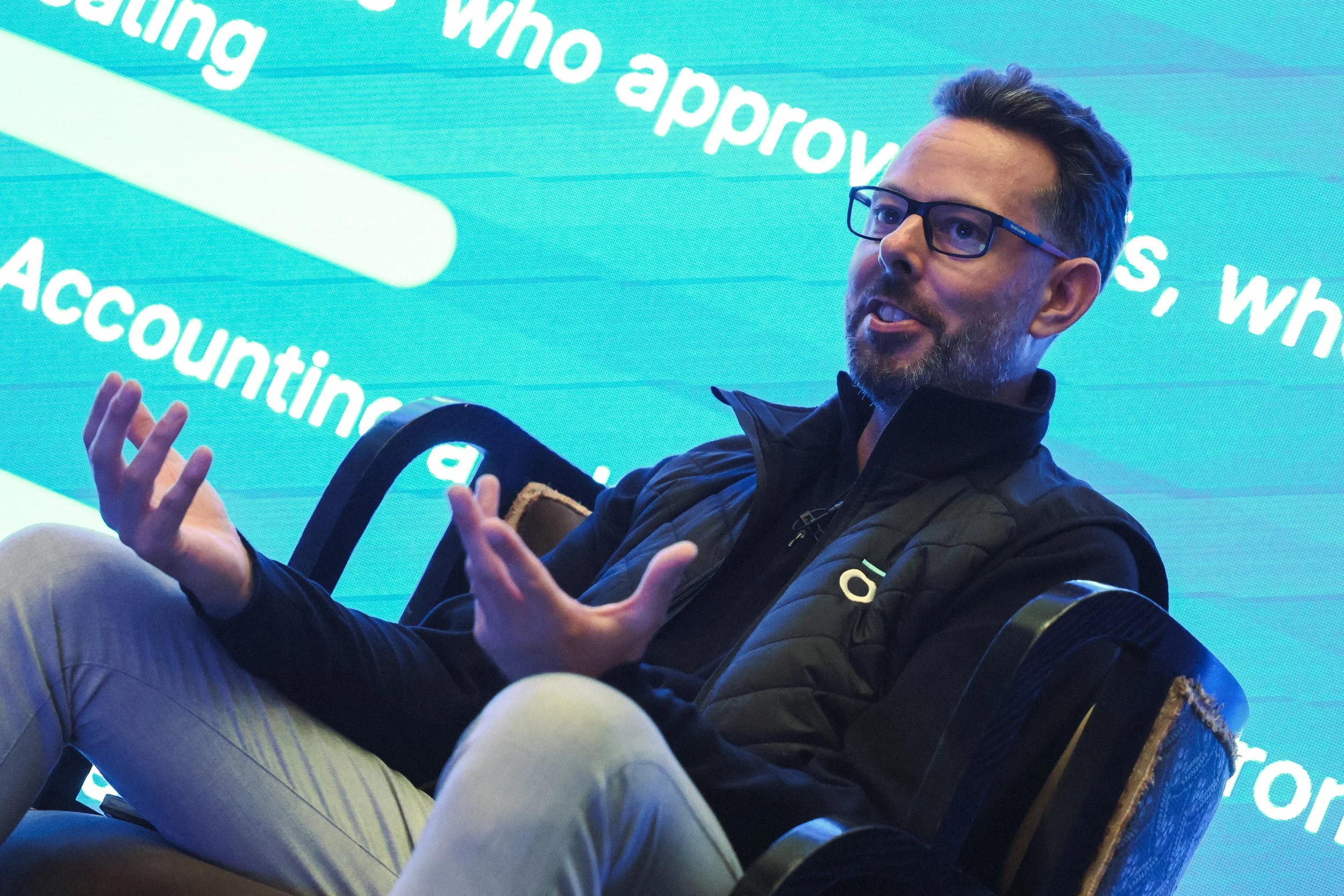 A man with glasses and a beard wearing a black jacket is sitting and speaking. There is a bright blue screen behind him with white text.