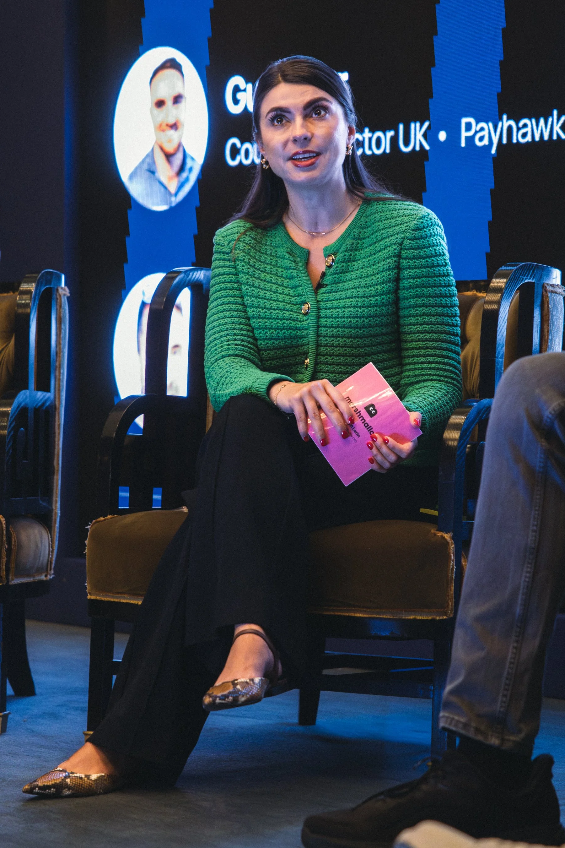 A woman with dark hair and fair skin is sitting in a conference or panel setting, holding a pink booklet in her hands. She is wearing a green textured jacket with gold buttons, black pants, and snakeskin-patterned shoes. She is speaking or listening 