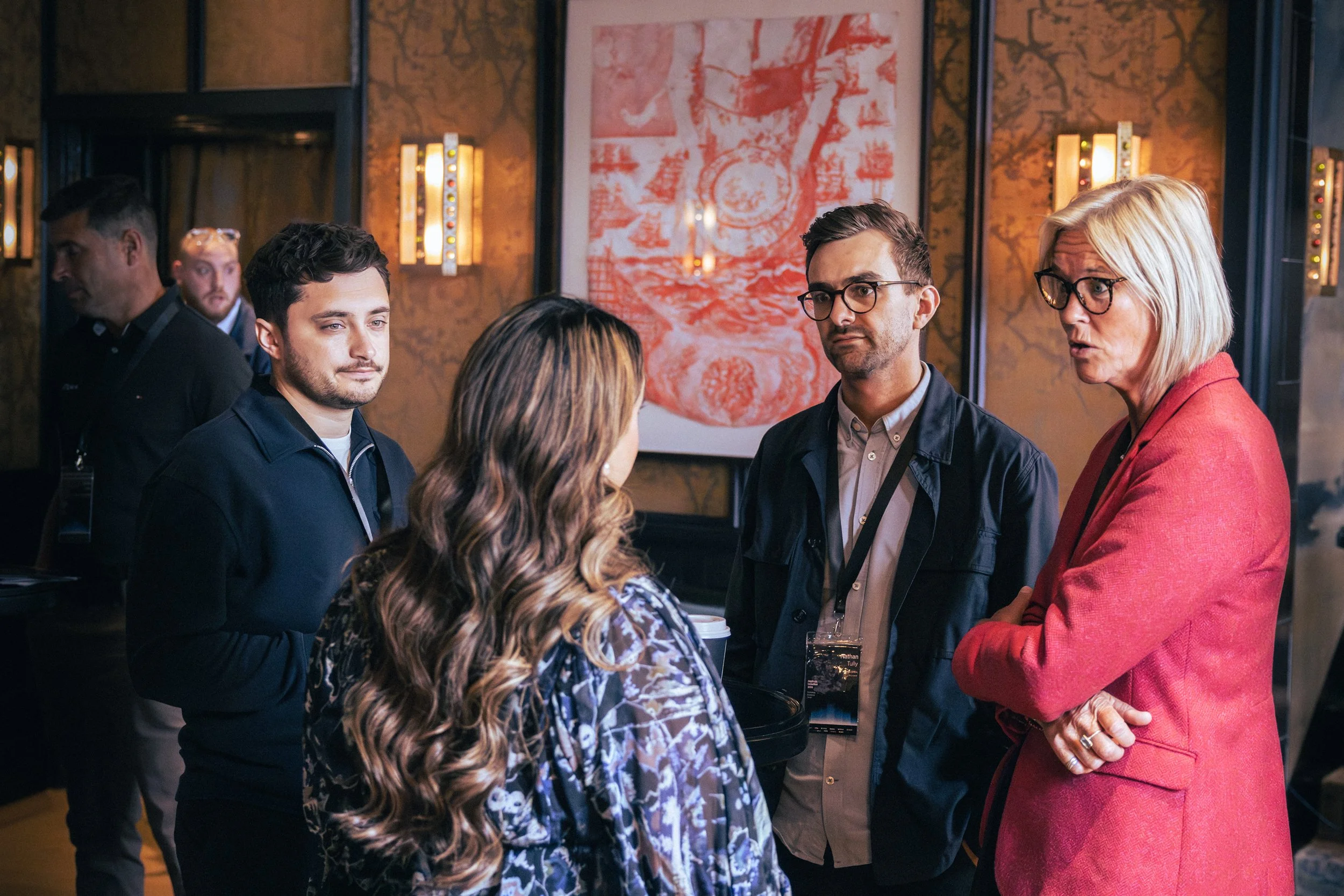 A group of people engaged in conversation at a social event inside a dimly lit room with artwork and wall-mounted lights in the background.
