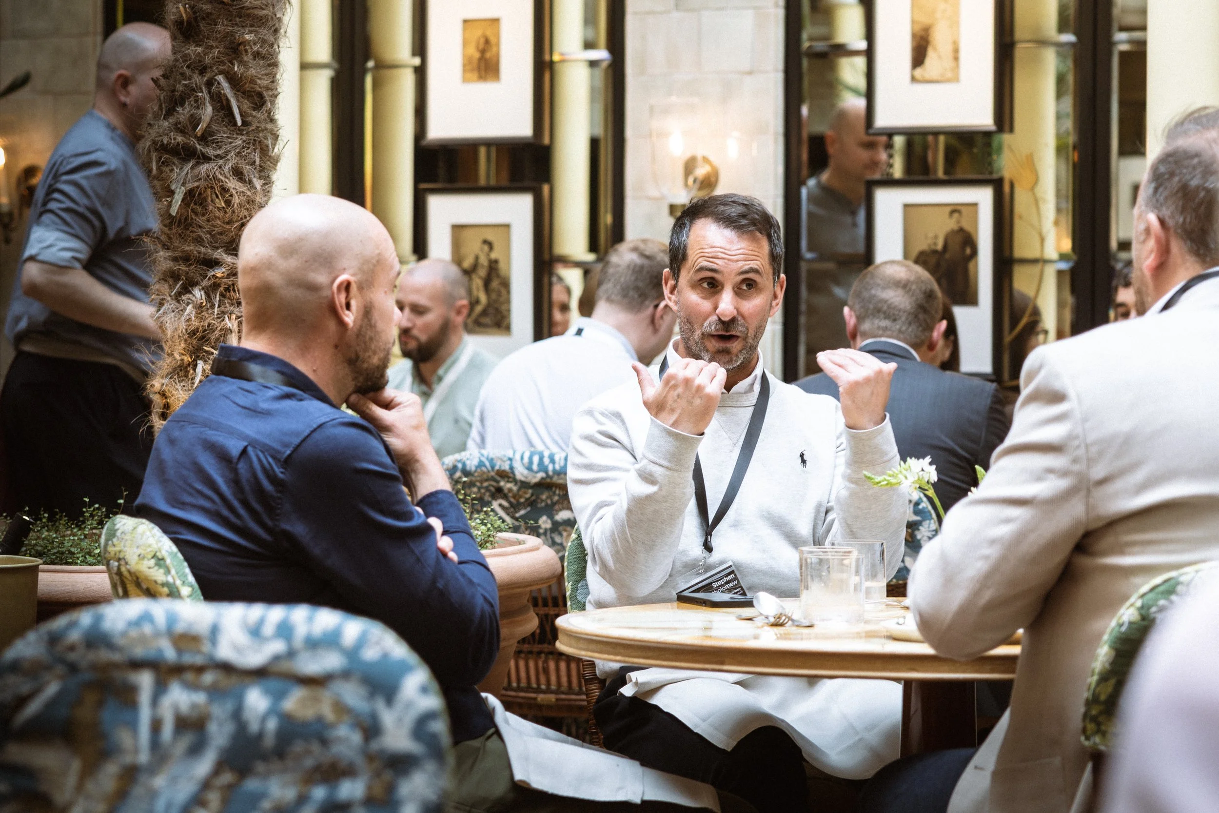 A group of people sitting around a table, engaged in conversation, with a man gesturing as he speaks. The setting appears to be a restaurant or a cafe with framed pictures and decorative objects on the wall behind them.