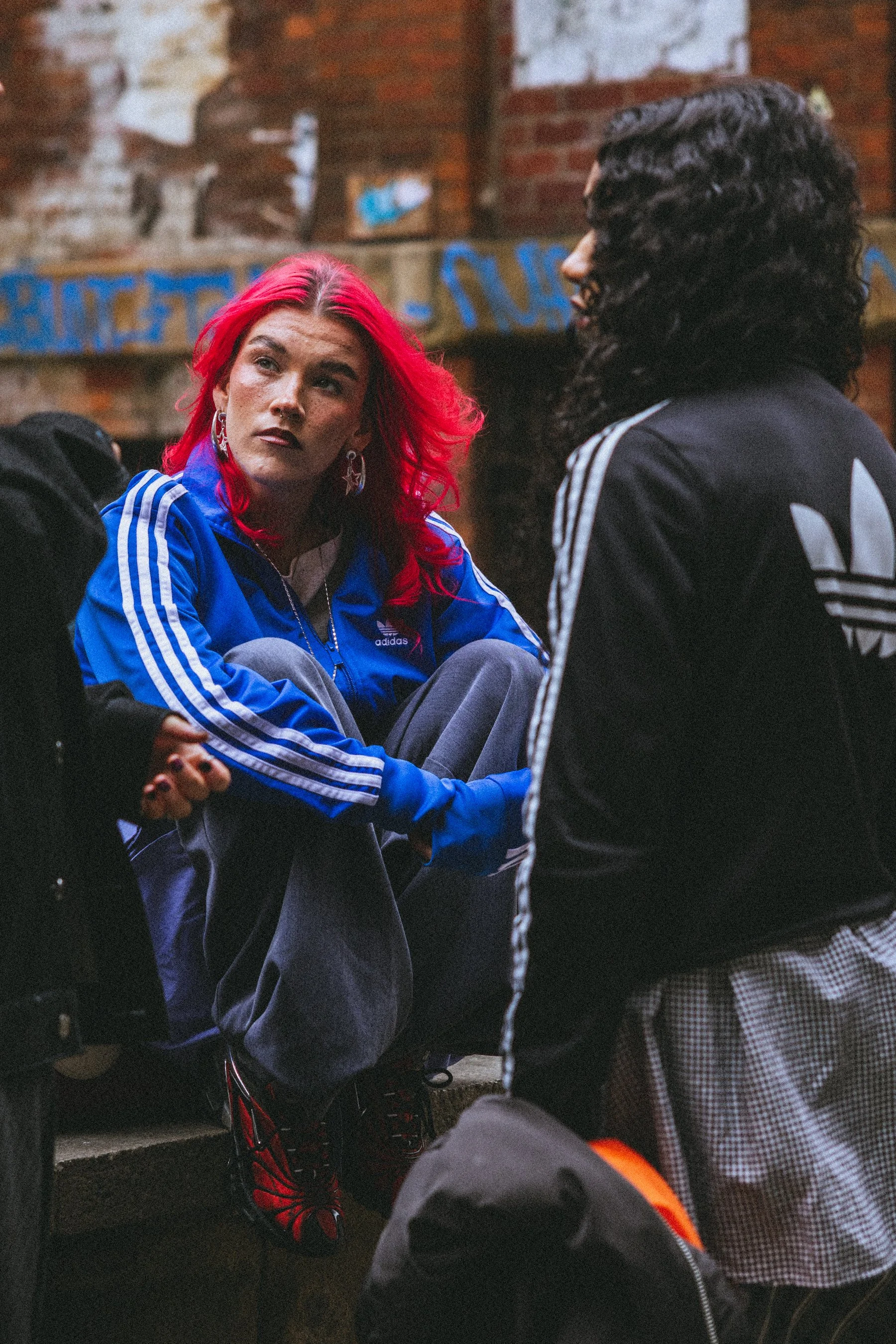 Two young people sitting together outdoors on a brick staircase. One has bright red hair and is wearing a blue Adidas jacket, while the other has black curly hair and is wearing a black Adidas jacket with white stripes.