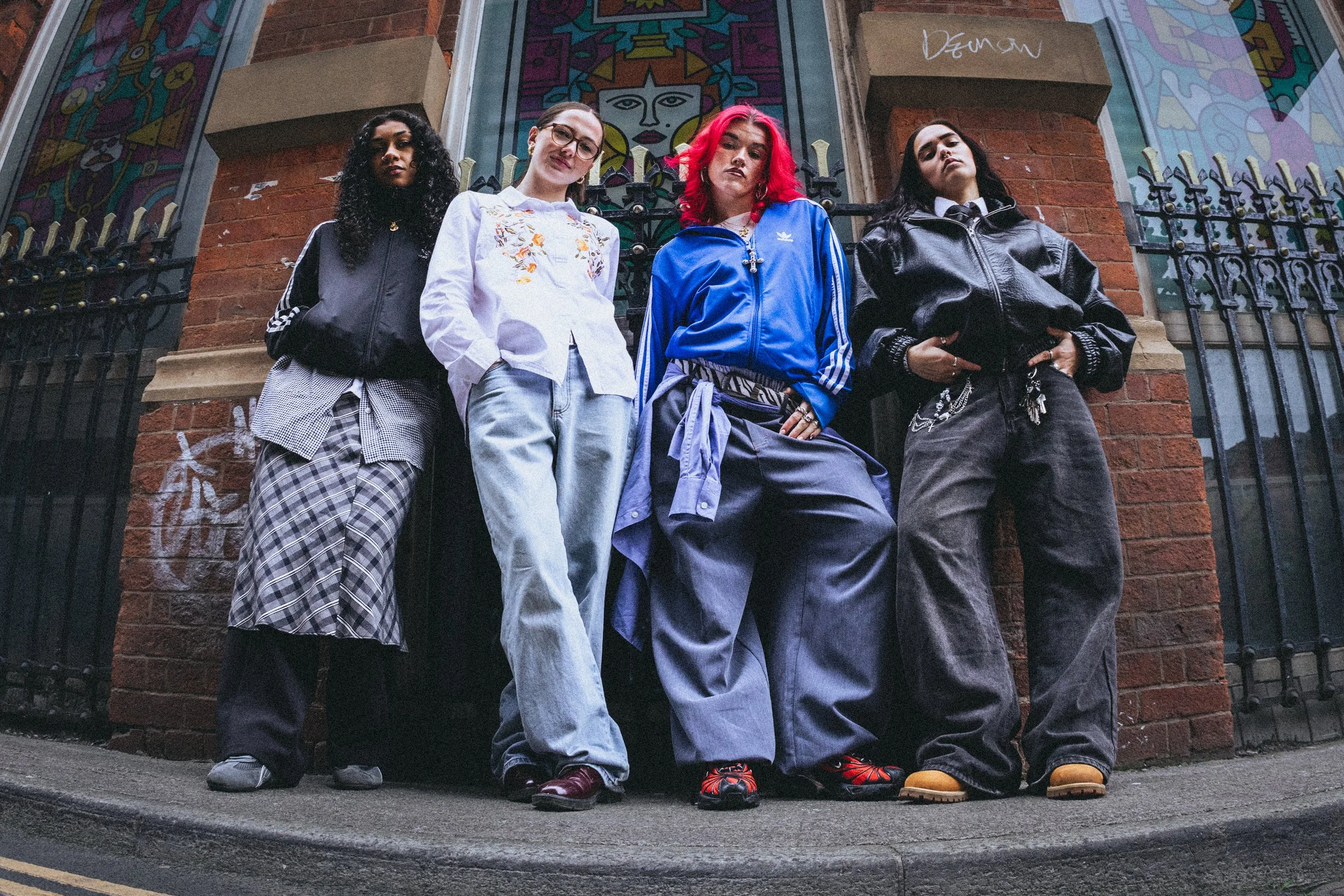 Four young people standing on a city pavement in front of a brick building with colourful artwork behind them. They wear casual streetwear, some with jackets and baggy pants, and have confident expressions.