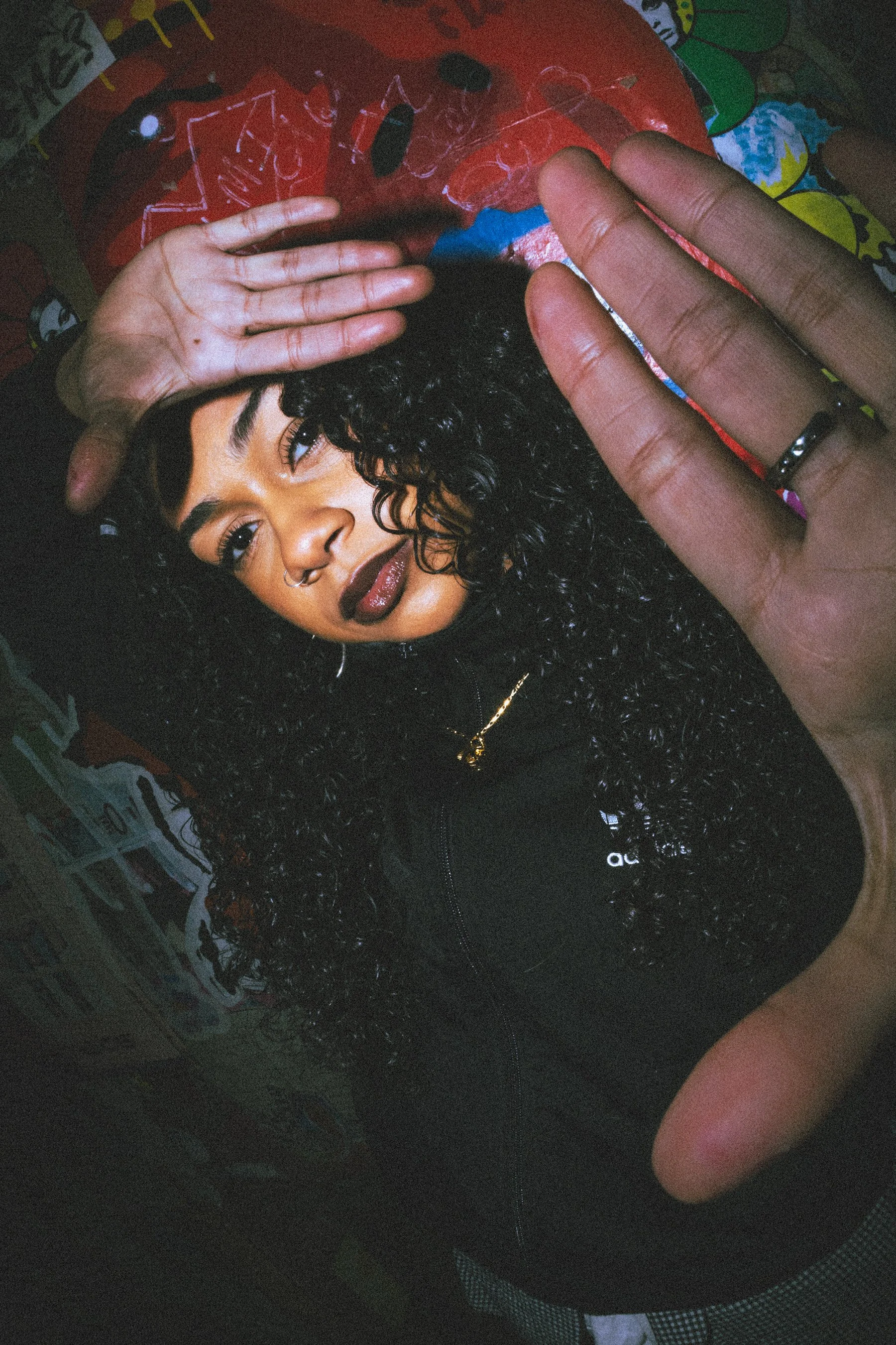 A person with curly black hair, wearing a nose ring, make-up, and a black jacket with a gold chain necklace, lying on a colourful background with her hand partially covering their face.