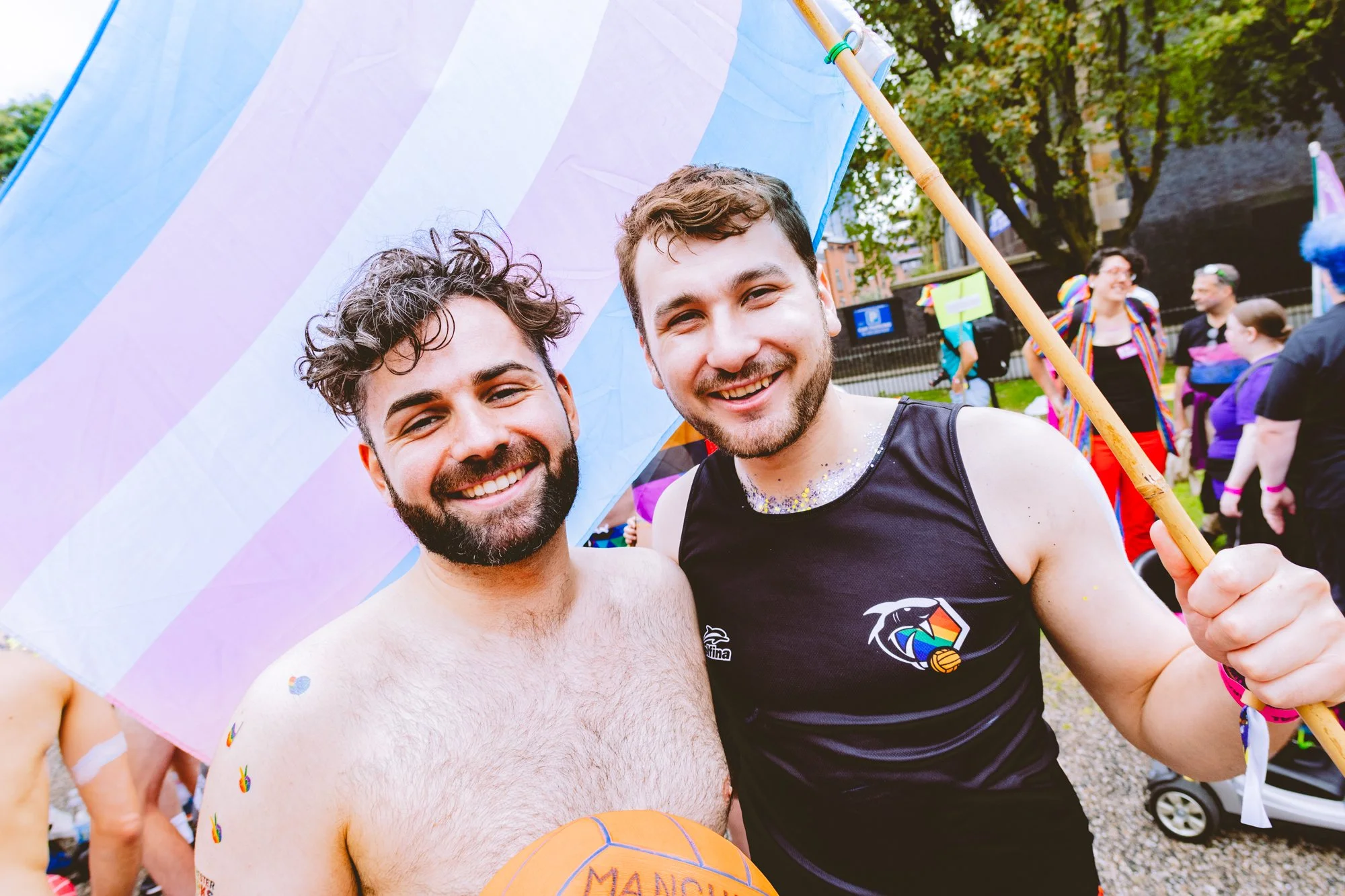 Two men smiling and posing for a photo at an outdoor LGBTQ pride event, one holding a rainbow umbrella and the other holding an orange basketball, with rainbow-themed clothing and colorful background.