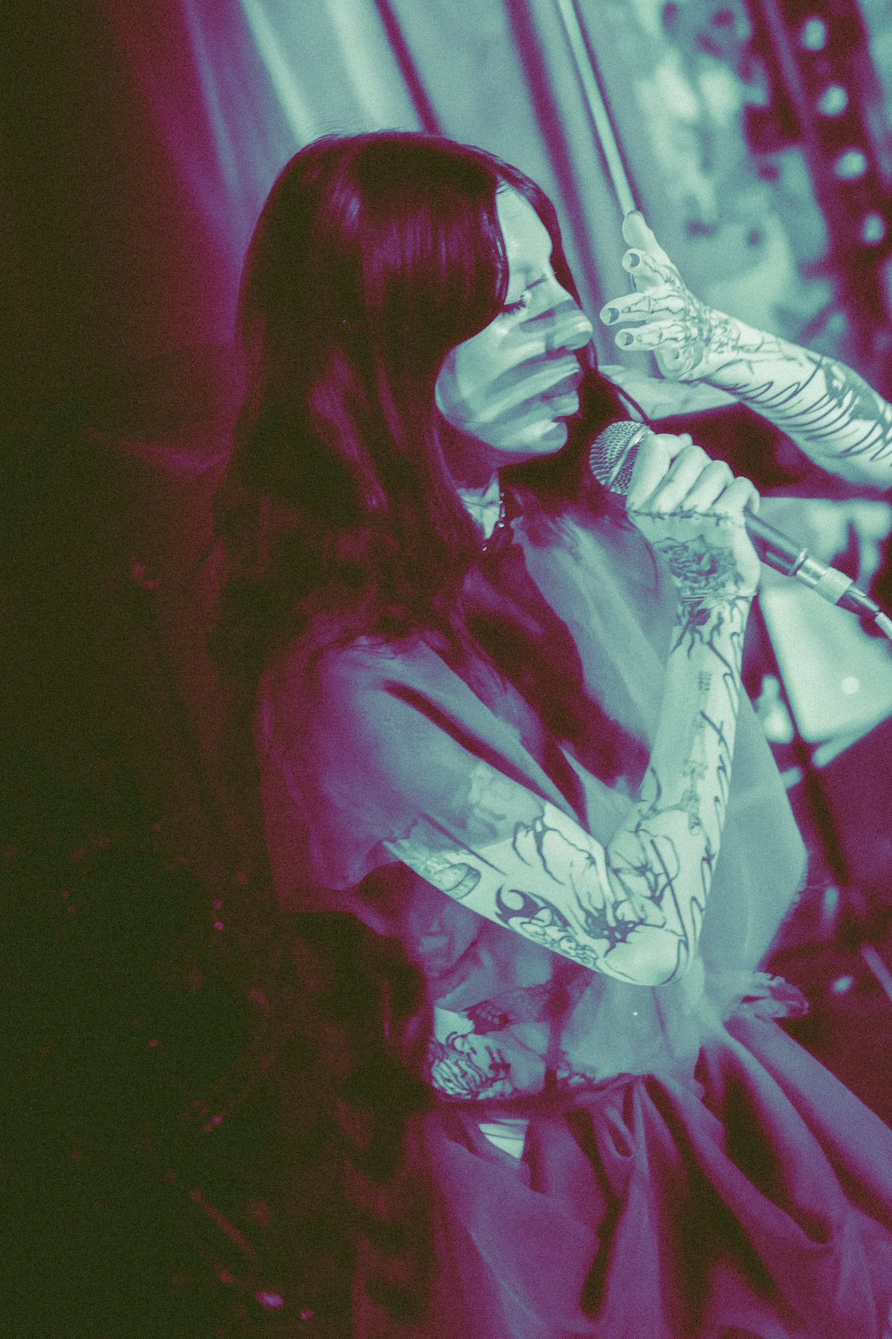 A performer with long dark hair and tattoos on their arms holding a microphone and singing with their eyes closed.