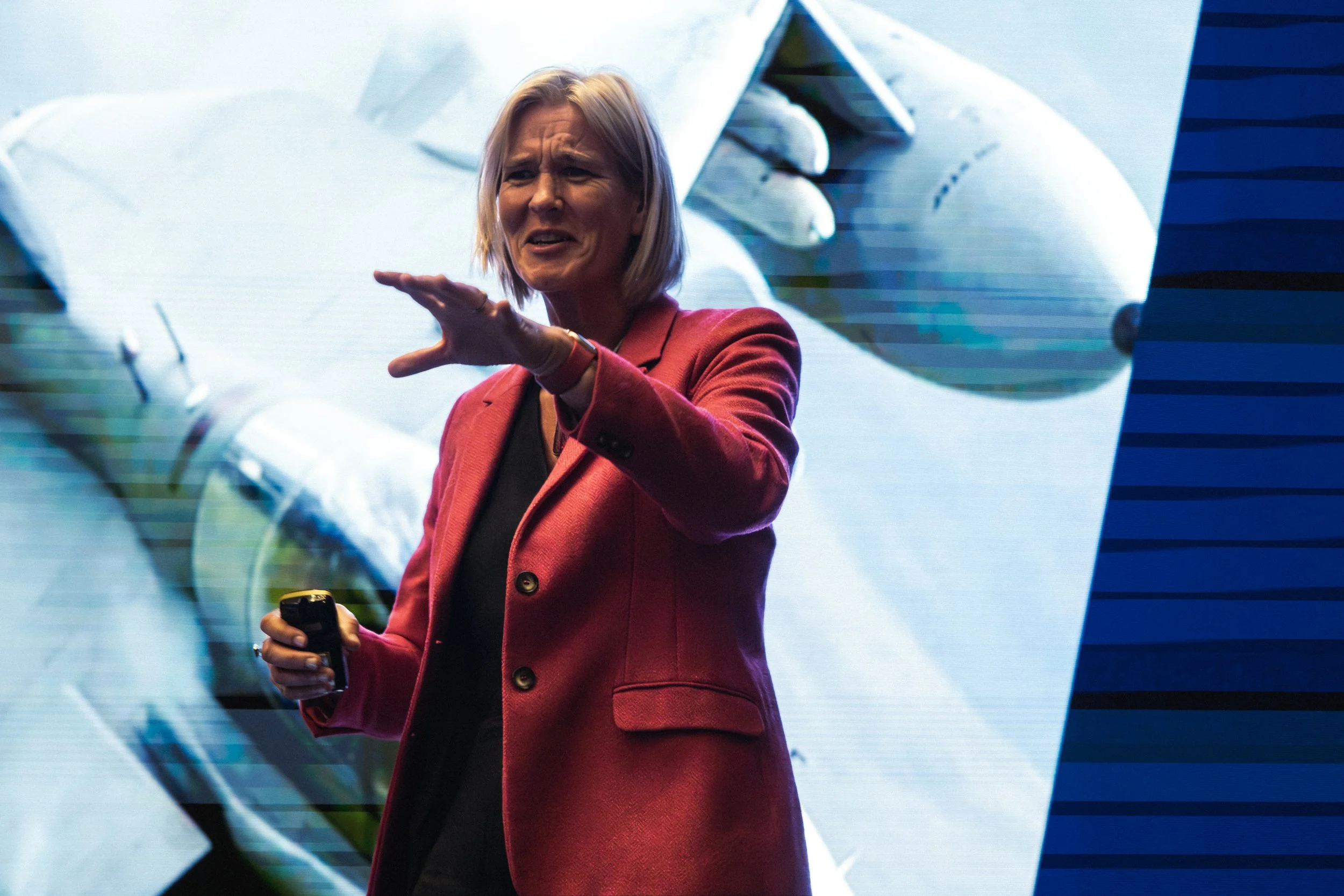 A woman with shoulder-length blonde hair wearing a red blazer giving a presentation or speech, holding a remote control, with a large screen behind her displaying blurred digital graphics.