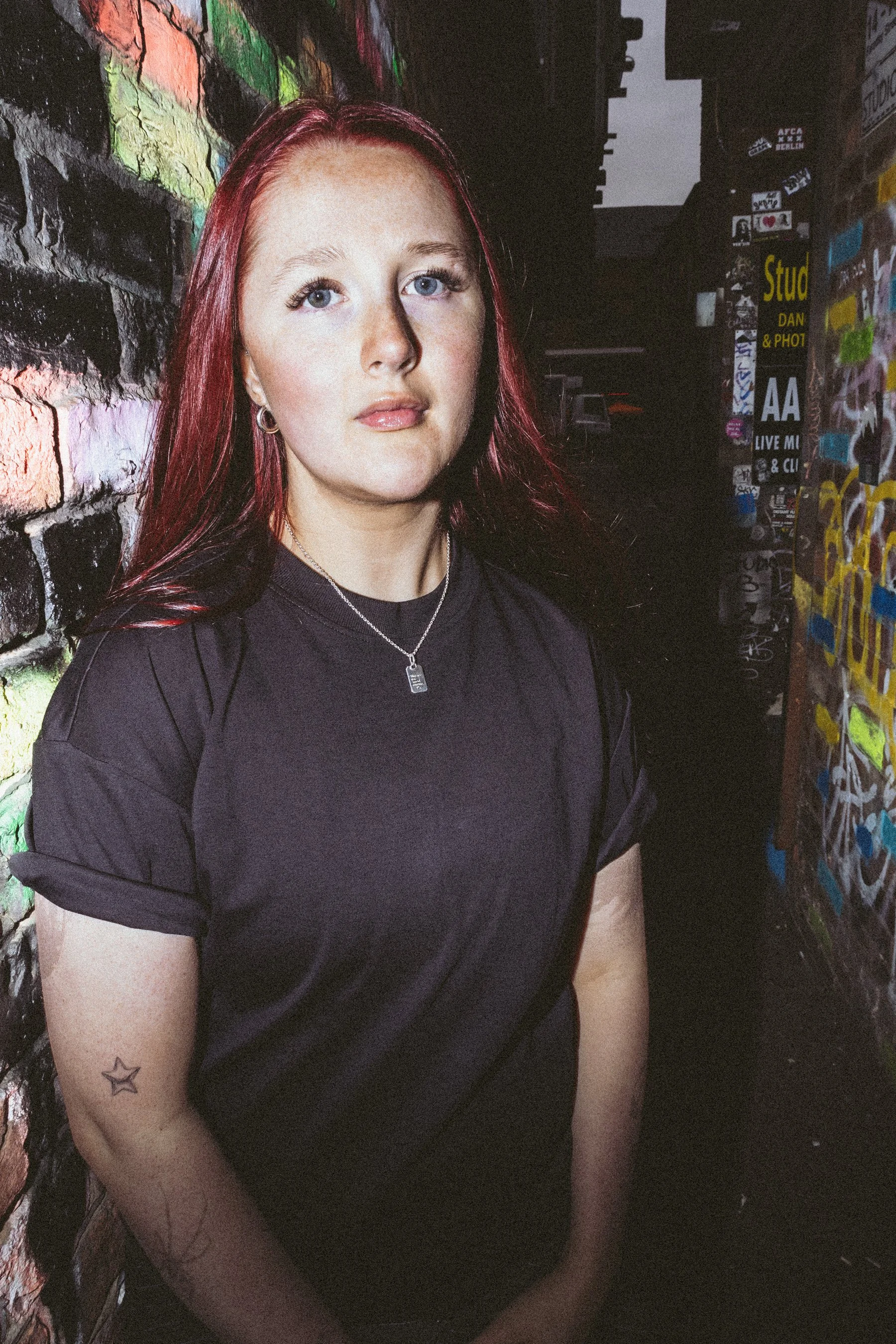 A woman with red hair and blue eyes stands in front of a graffiti-covered brick wall and a wall with stickers and posters, with a dark alleyway behind her.