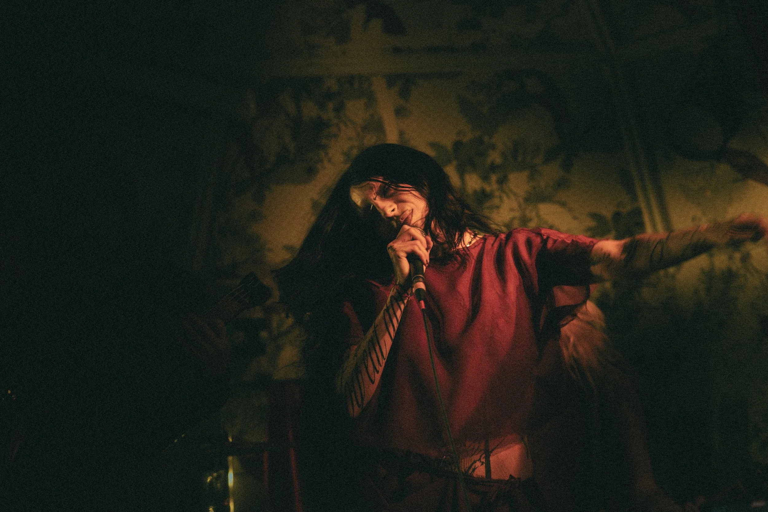 A singer with long dark hair singing into a microphone on stage, wearing a red shirt.