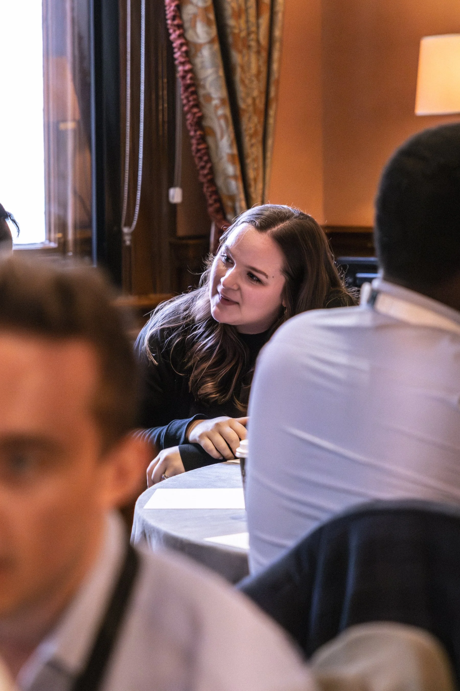Young woman with long brown hair engaged in conversation at a social gathering inside a warmly lit room, with ornate curtains and a table in front of her.