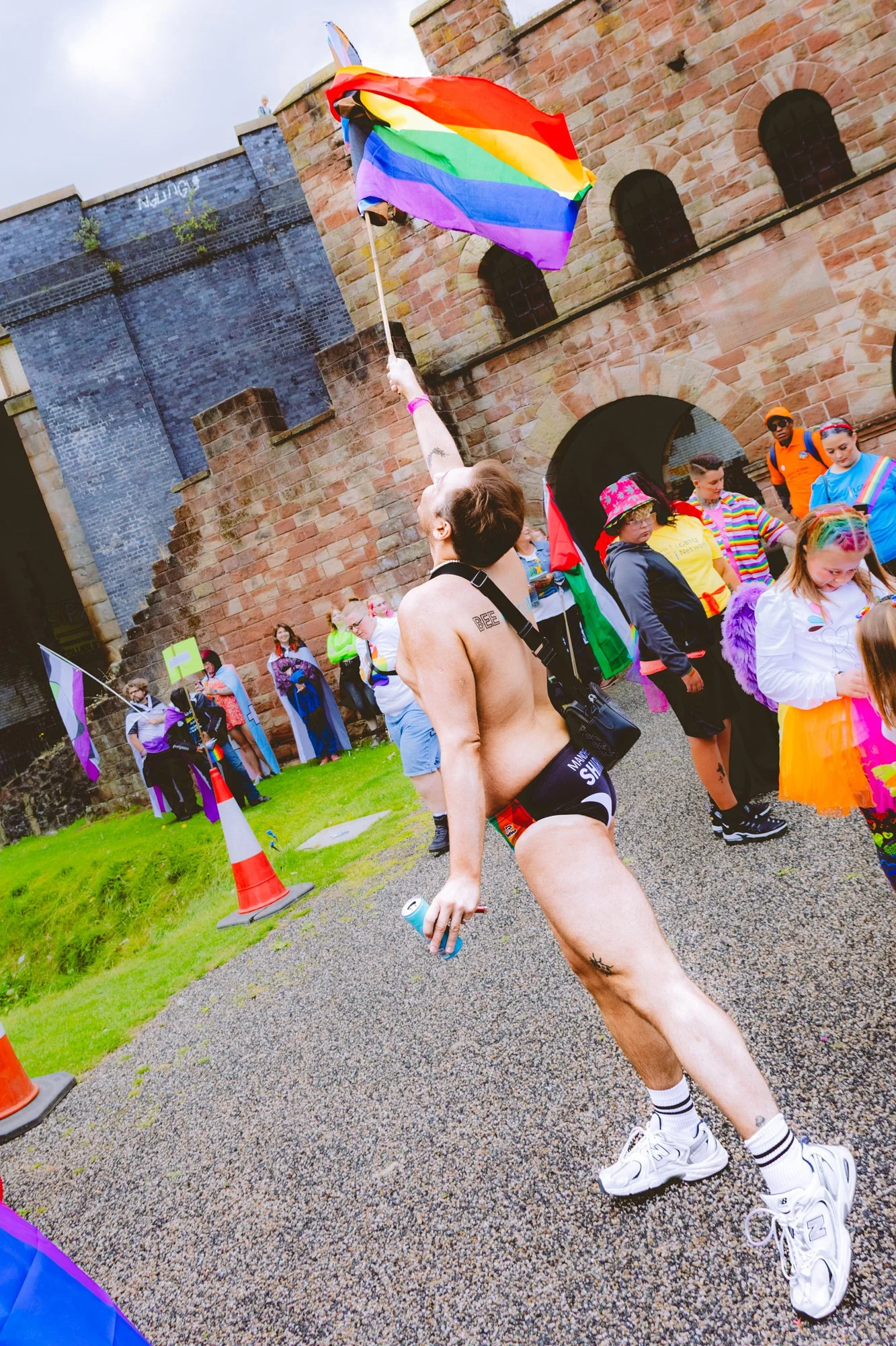 Person dressed in underwear holding a rainbow flag at a pride event, with a crowd and historic brick buildings in the background.