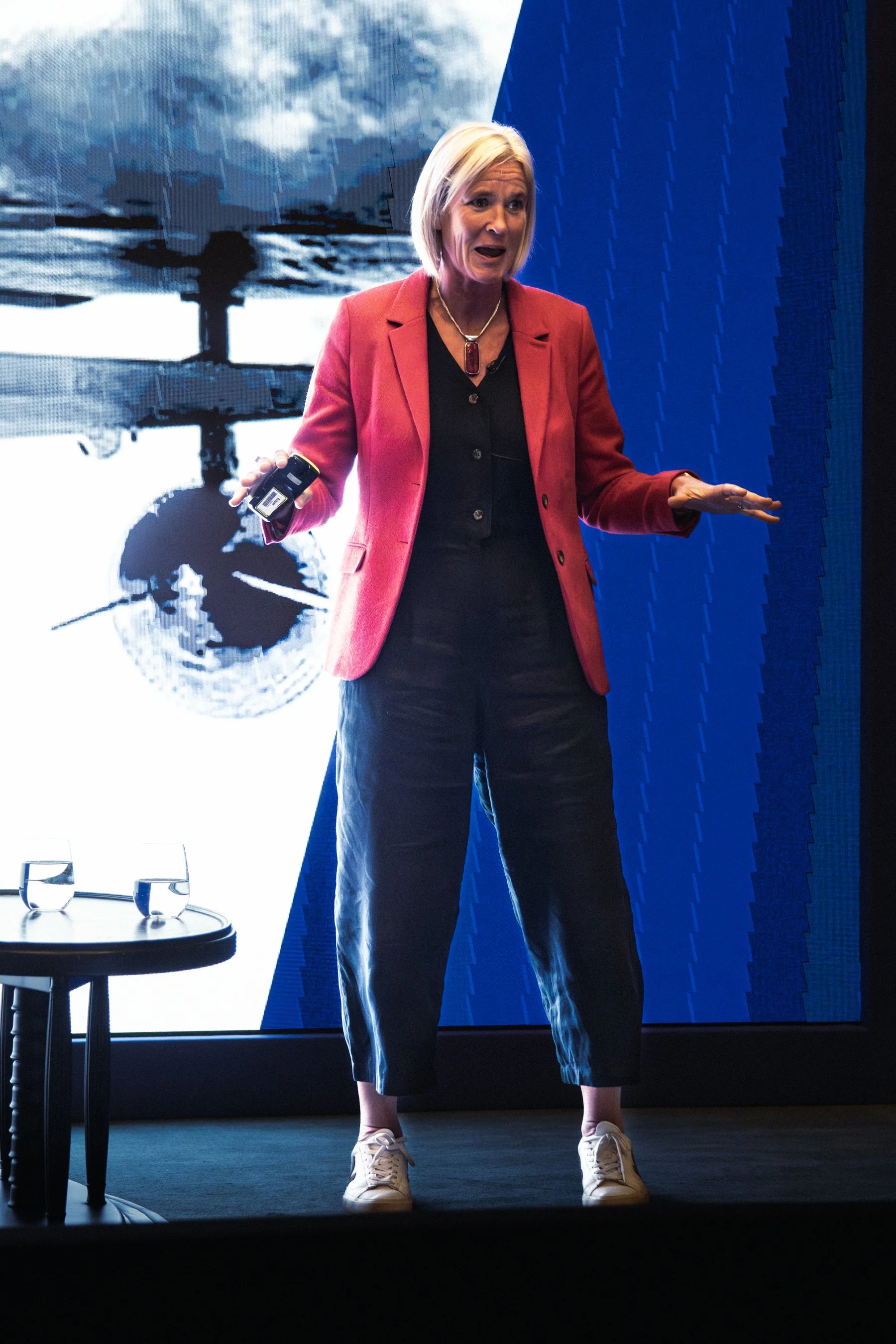Woman in a red blazer and black pants giving a presentation, standing in front of a large screen at a conference.