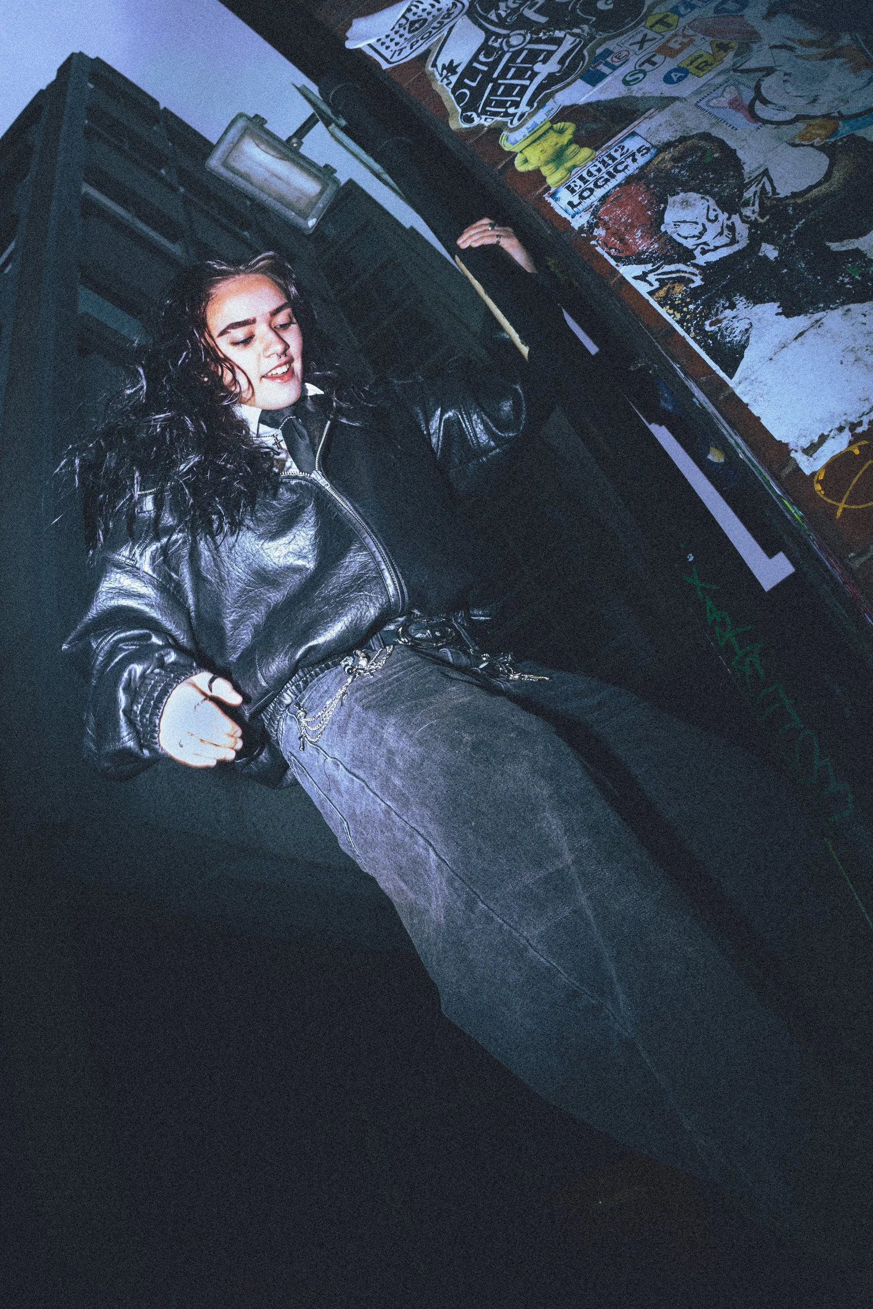 A young person with long, dark hair, wearing a black leather jacket and jeans, standing half way up a lamp post.
