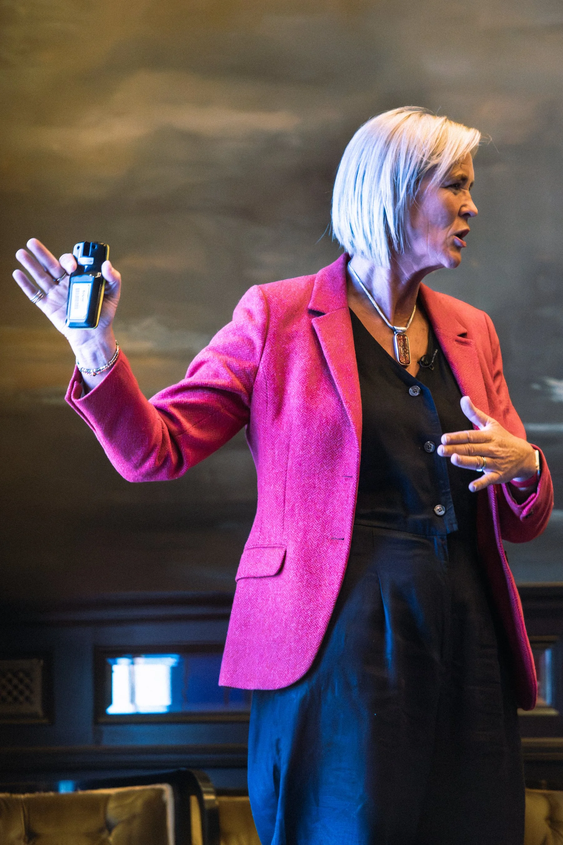 A woman with shoulder-length blonde hair wearing a pink blazer and black top, speaking passionately while holding a remote or phone in her right hand at an event.