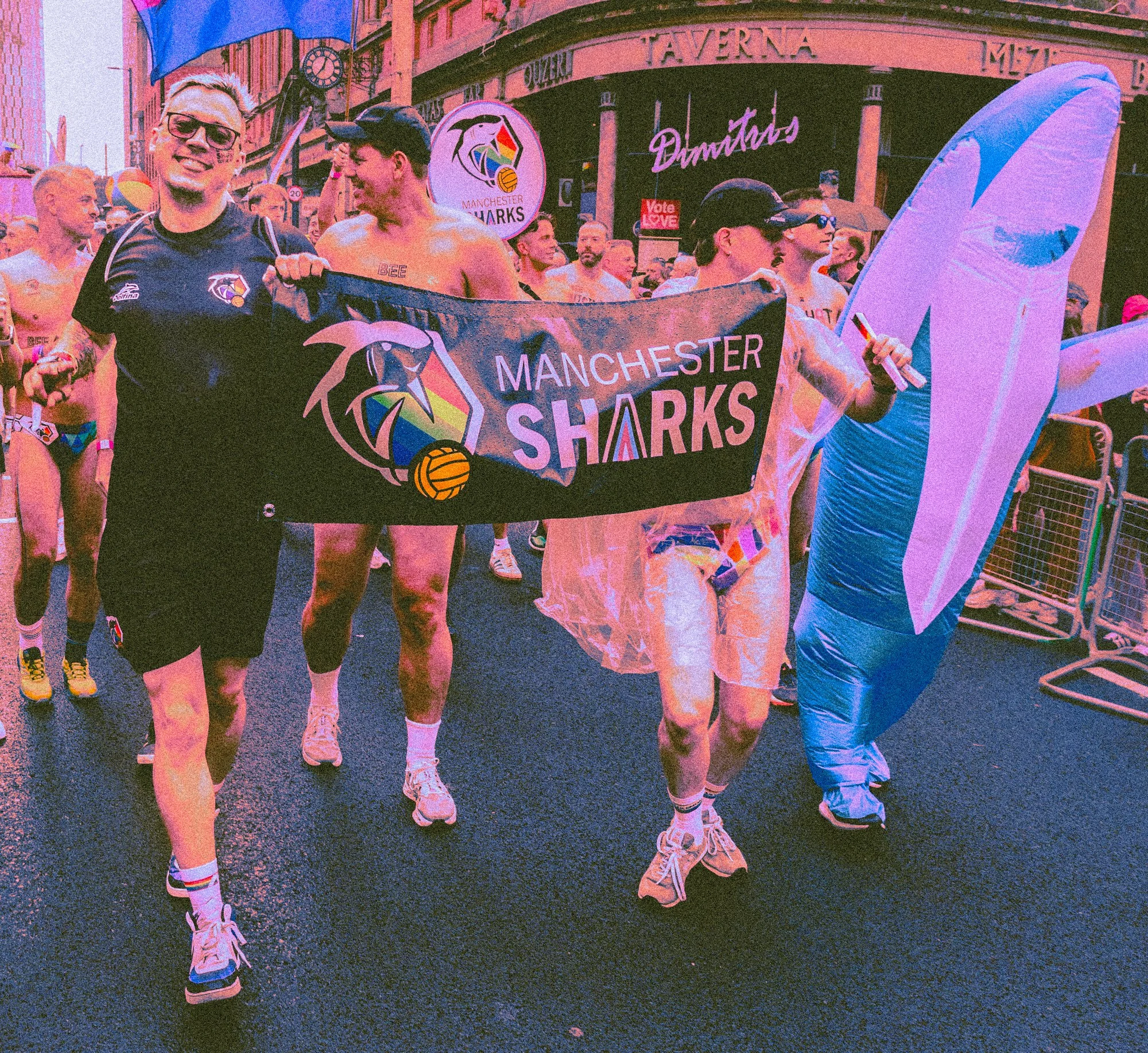 A group of people participating in a Pride parade. Some are wearing sports jerseys and shorts, walking on a city street. One person is holding a Manchester Sharks banner, and another is holding a rainbow flag. The background shows buildings and signa