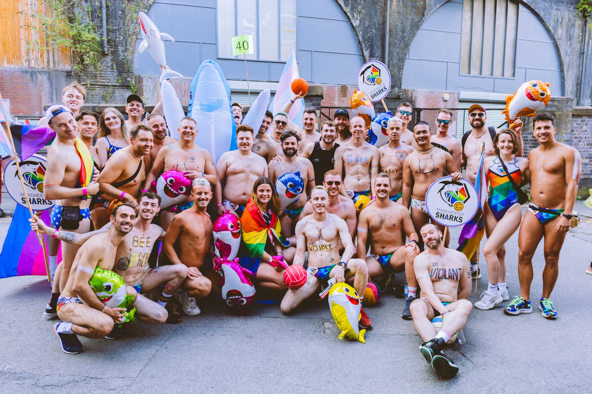 Group of people in swimwear holding balloons with sharks and rainbow flags, celebrating Pride month, outdoors near an urban background with brick walls and an arched structure.