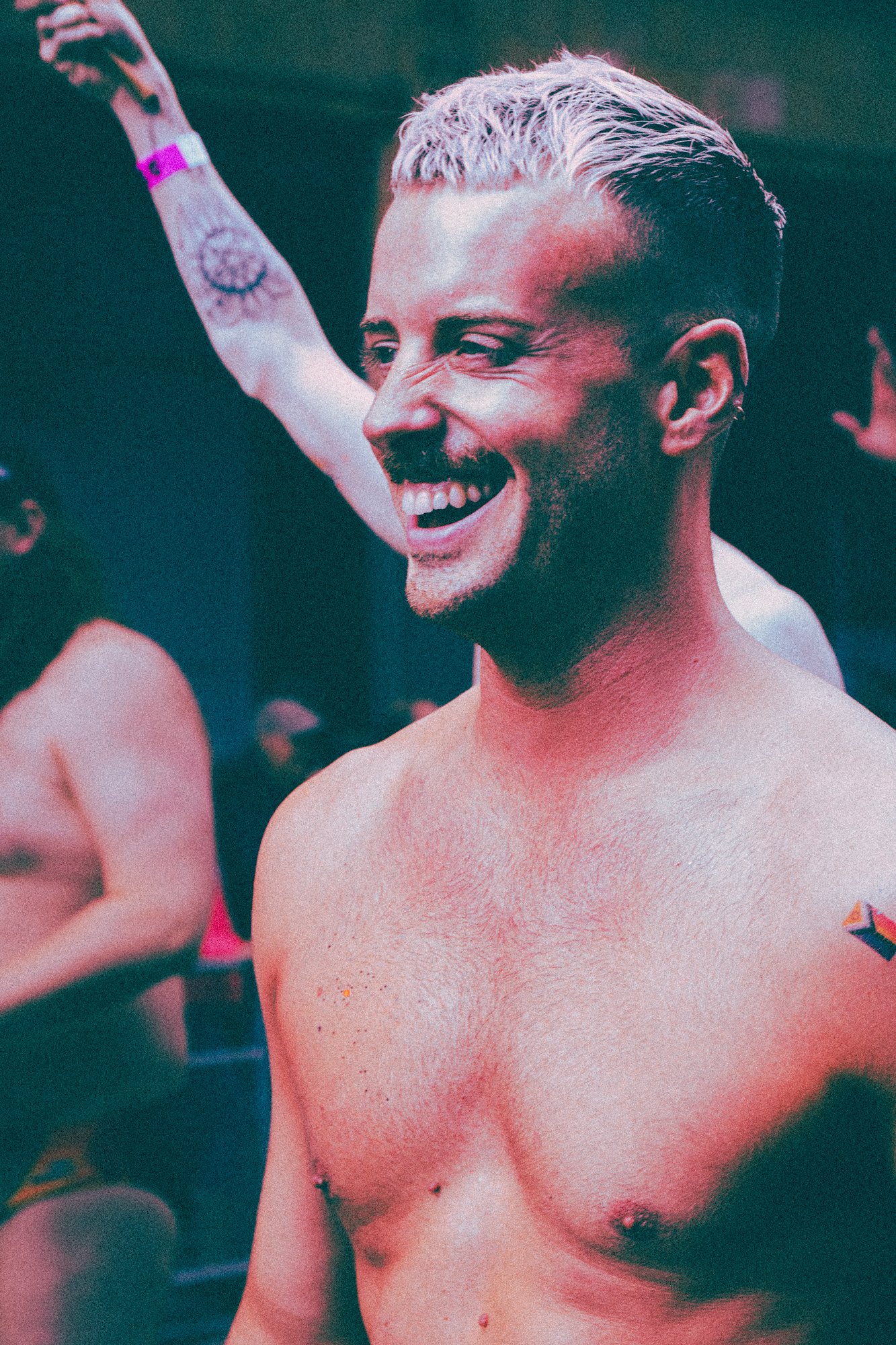 A shirtless man with platinum blonde hair smiling at an event or gathering, with other people visible in the background.