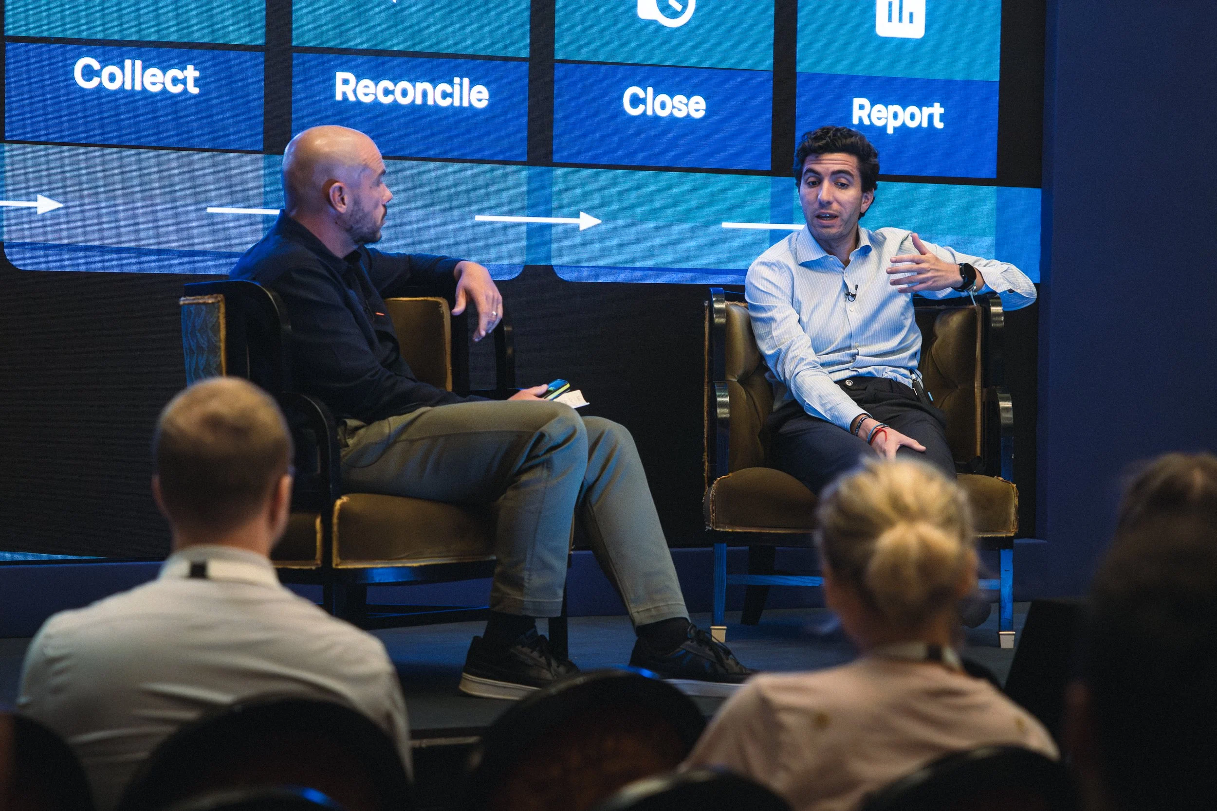 Two men are seated on stage in front of a large digital screen during a presentation or discussion. The screen displays words like 'Collect,' 'Reconcile,' 'Close,' and 'Report,' with arrows connecting the steps. The audience's heads are visible in th