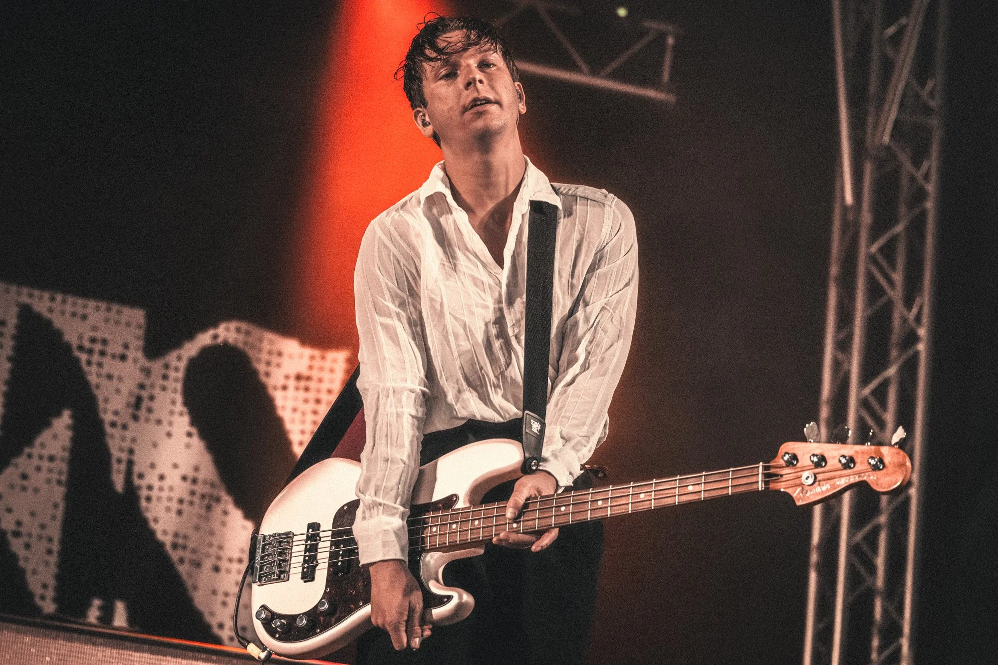 A musician playing an electric bass guitar on stage, wearing a white striped shirt and ear monitors, with stage lighting and a backdrop in the background.