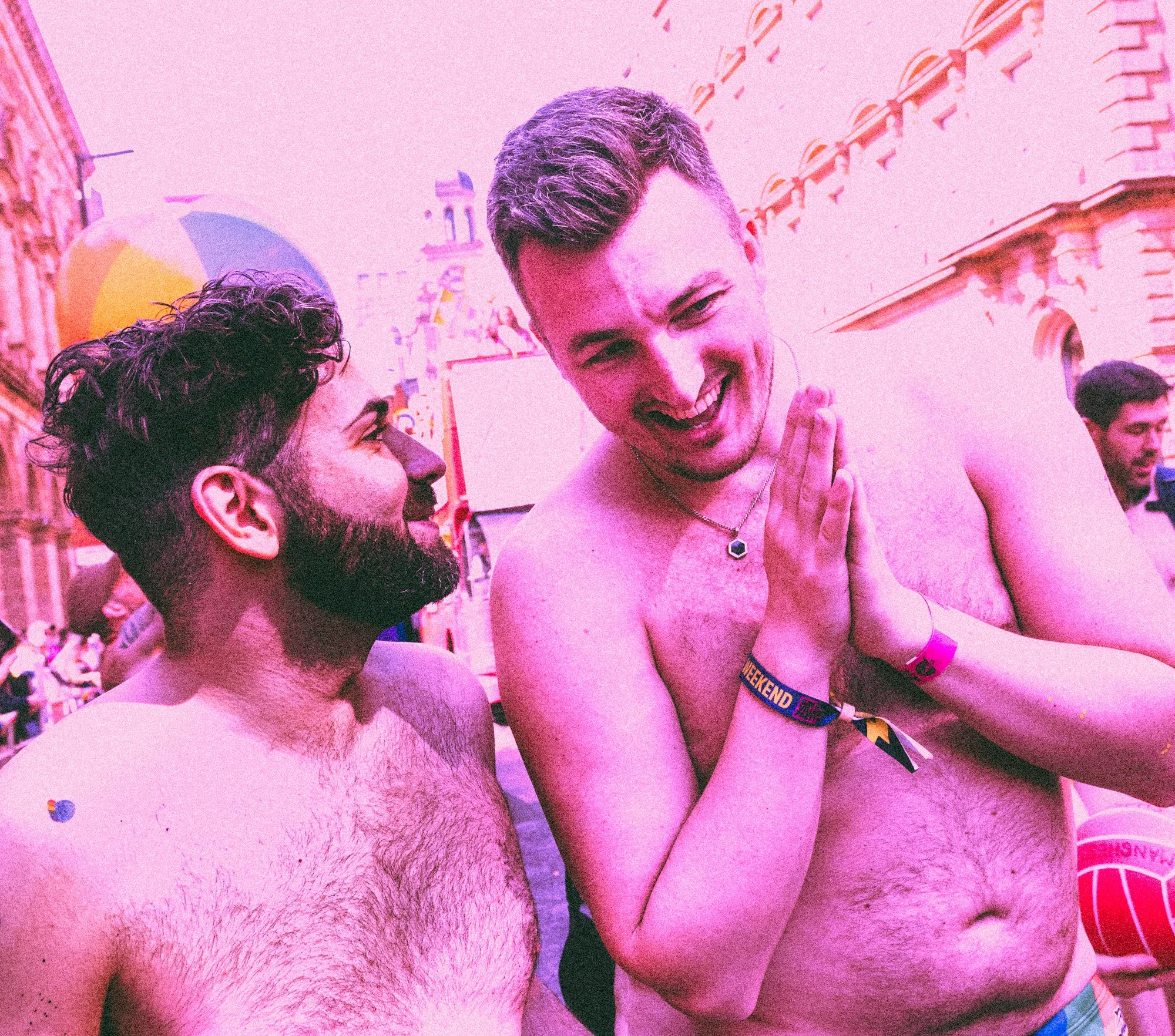 Two shirtless men with beards conversing at Manchester Pride with a pink-colored filter, with buildings in the background.