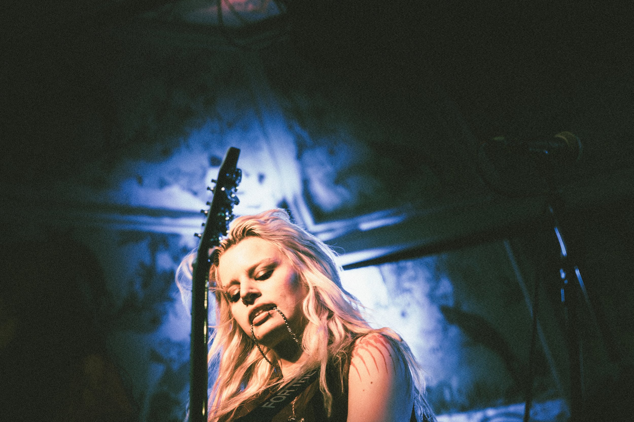 A woman with long blonde hair and tattoos has her head slightly lowered with eyes closed, holding a black chain in her mouth, in a dimly lit room with blue light and a microphone stand in the background.