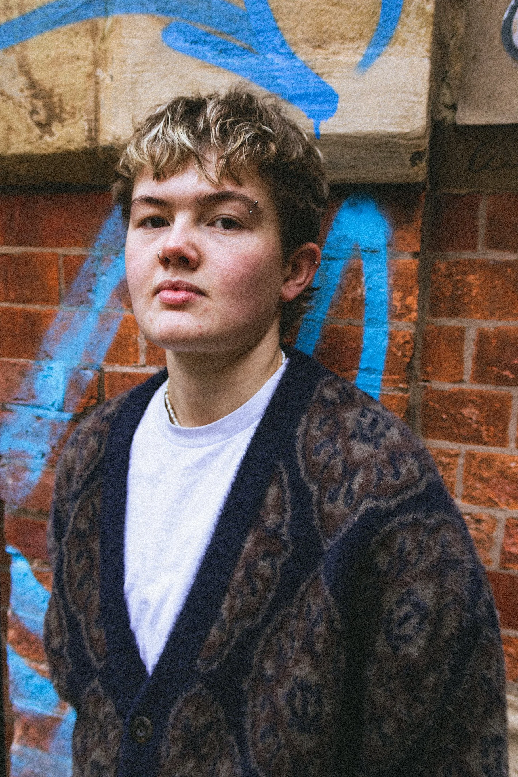 Young person with short, curly hair and facial piercings, wearing a leopard-print cardigan over a white t-shirt, standing against a brick wall with blue graffiti.