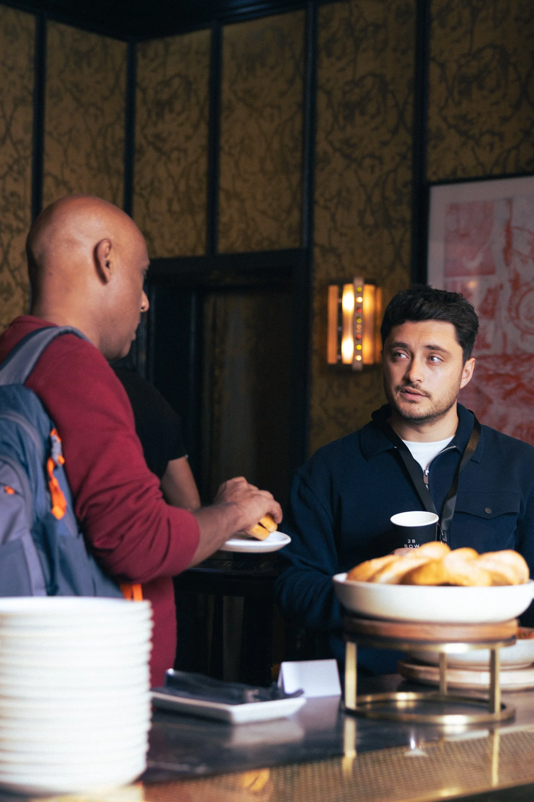 Two men are having a conversation at a buffet table with plates of food, including a bowl of bread rolls, visible in the foreground. One man with a bald head and backpack wears a red shirt, and the other man with dark hair in a dark jacket holds a bl