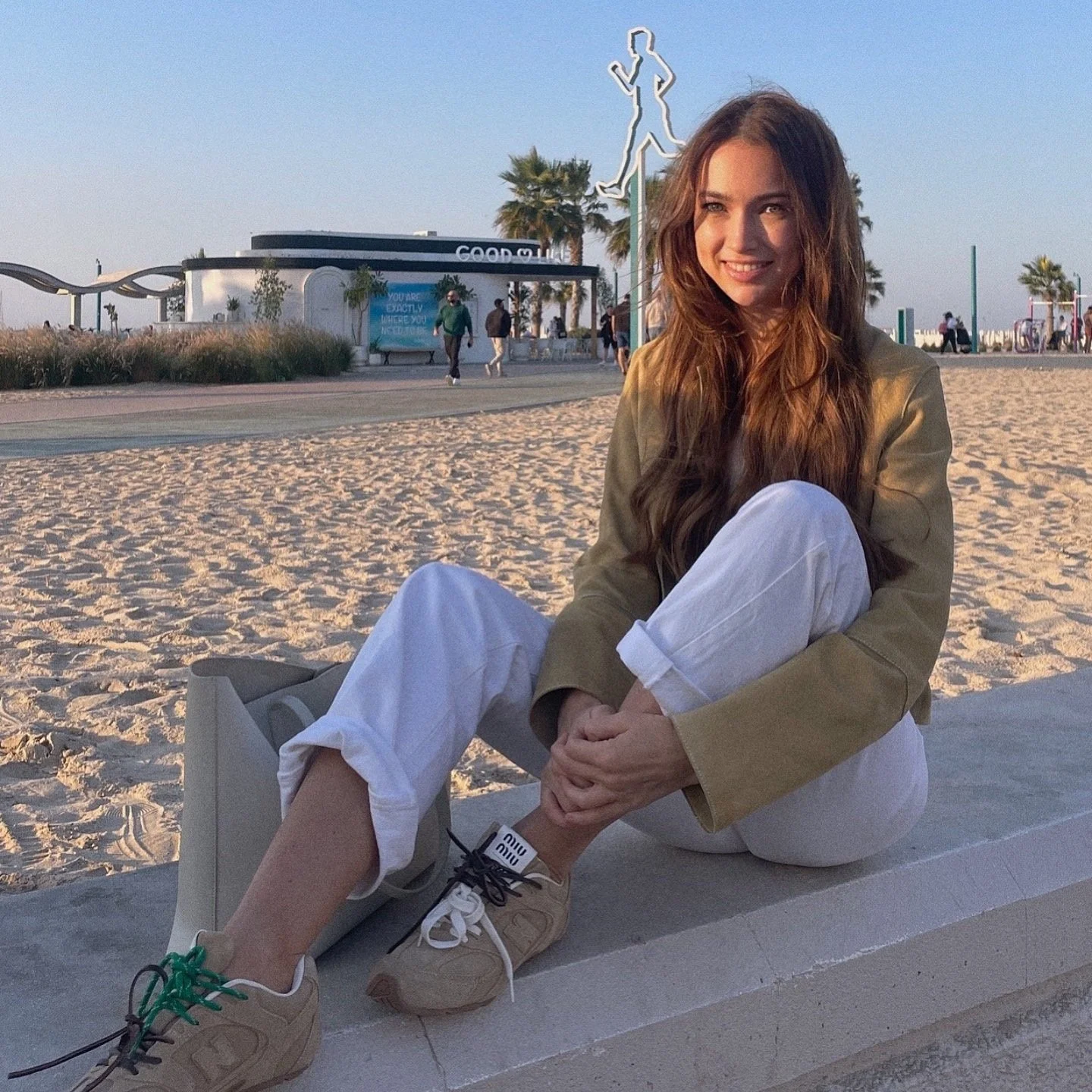 A woman sitting on a sandy beach, wearing a beige jacket and white pants, with green and black shoelaces on tan sneakers ( MiU MIU x New Balance). A background of palm trees and a café with an outdoor seating area is visible.