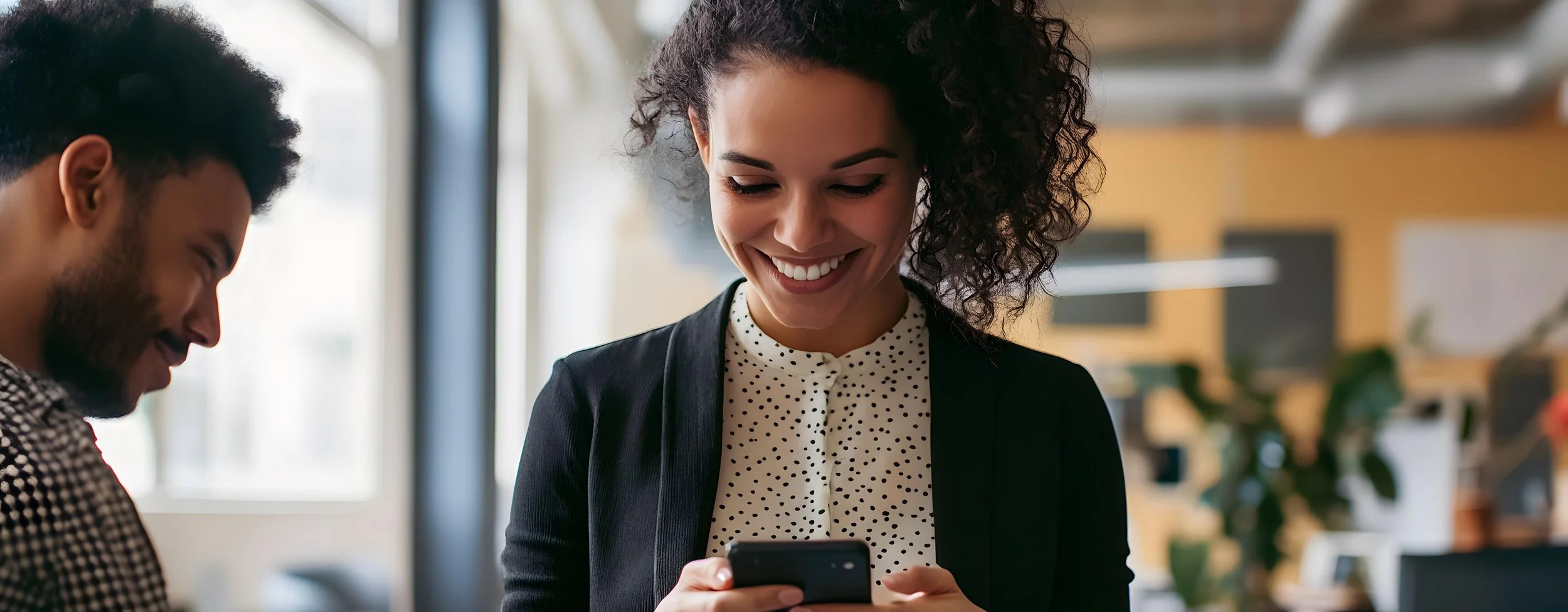 Two people, a woman and a man, smiling and looking at a smartphone in a modern office space.